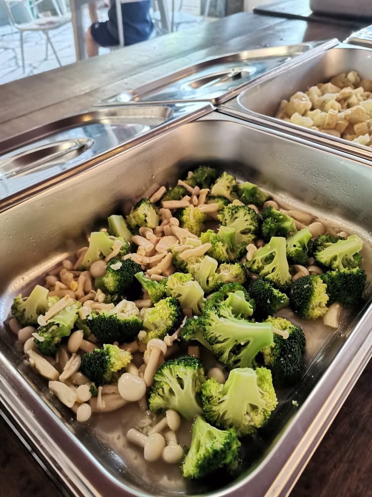 Steamed broccoli and shooting stars mushrooms in a metal serving tray at a buffet.