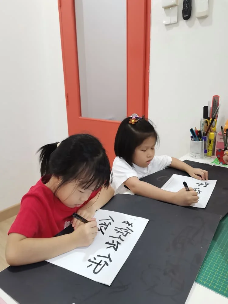 Two young girls practicing calligraphy or writing Chinese characters on white paper at a black table with various art supplies in a room.