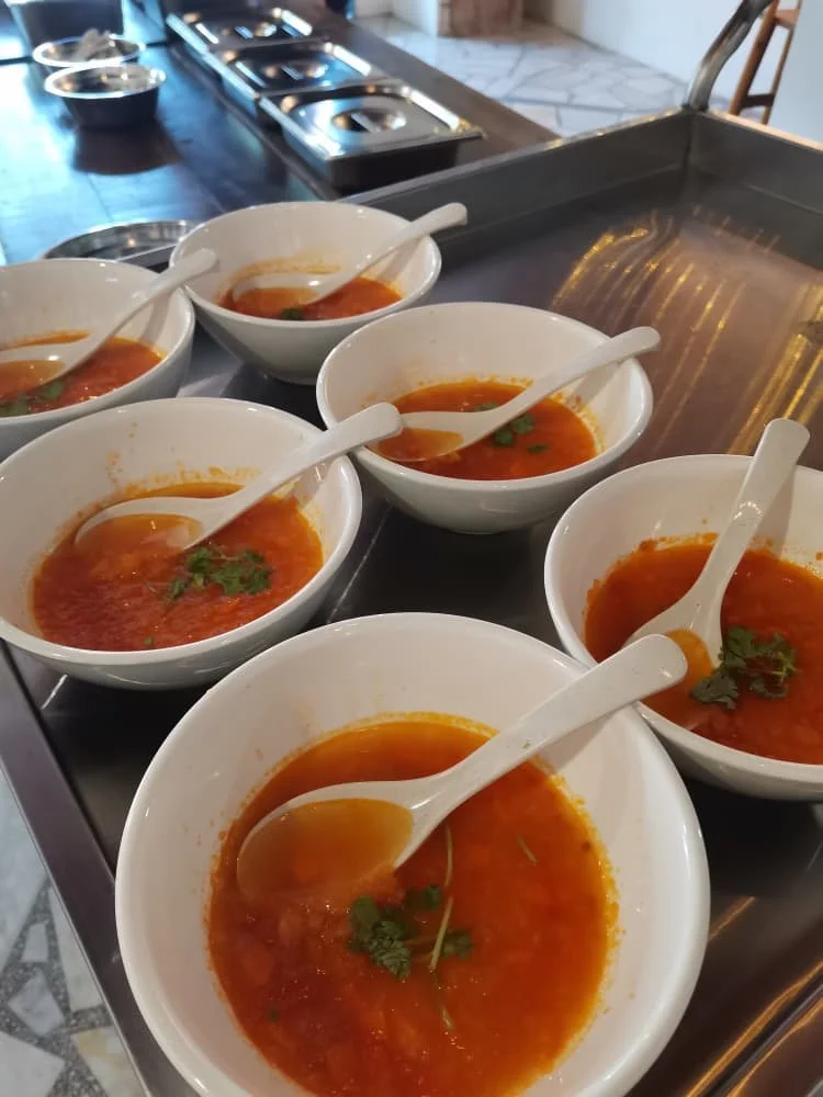 Six bowls of tomato-based soup with herbs, each with a white spoon inside, on a kitchen counter.