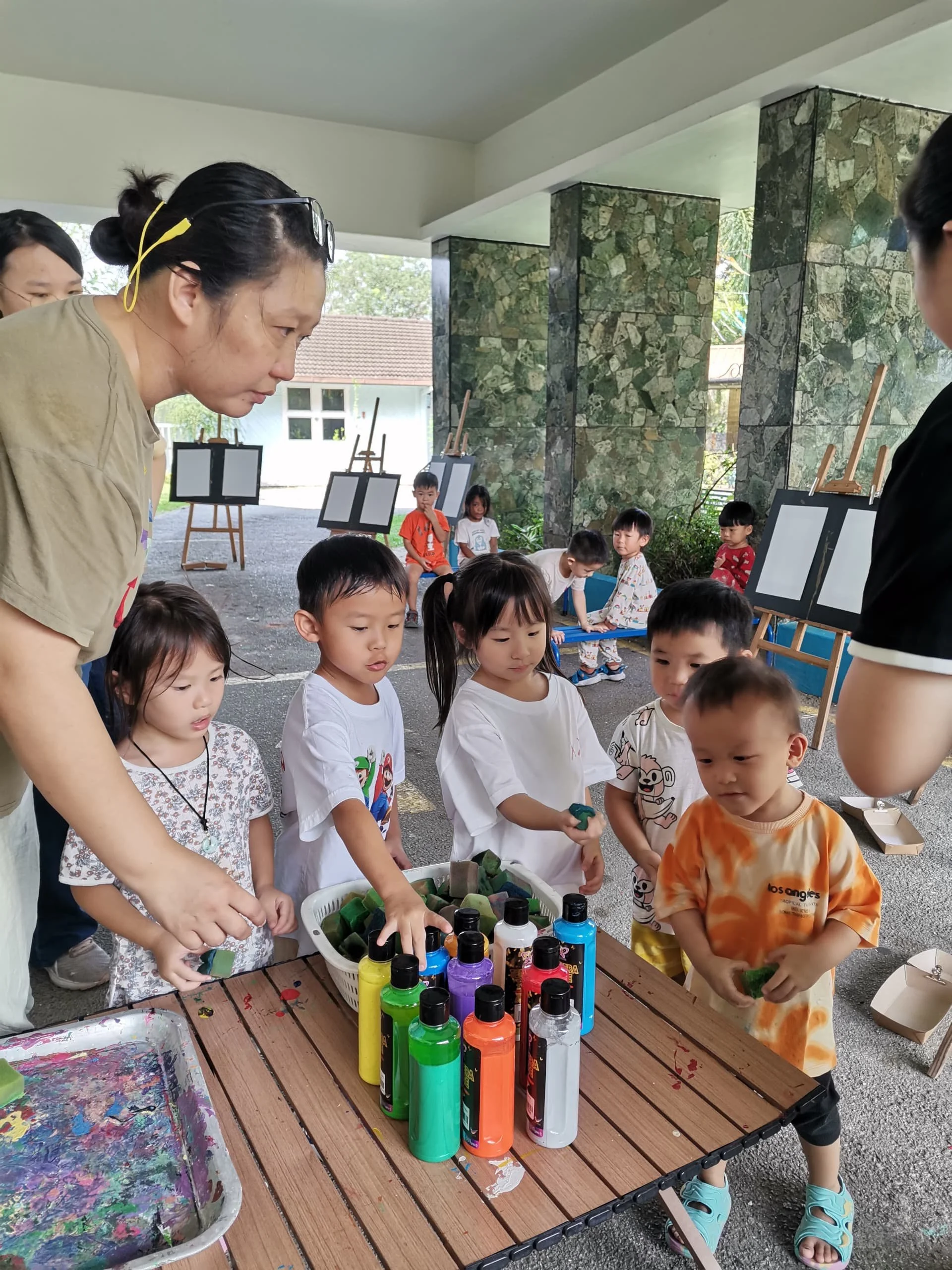 Children and adults participating in a painting activity outdoors, with paint bottles and art supplies on a wooden table, and easels with blank canvases in the background.