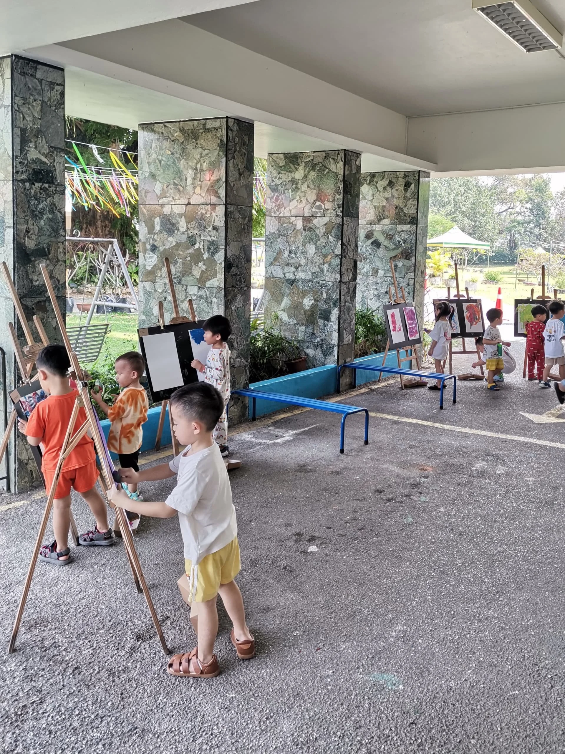 Children participating in an outdoor art activity, painting on easels under a shelter with stone pillars and a gravel ground, with trees and a tent visible in the background.