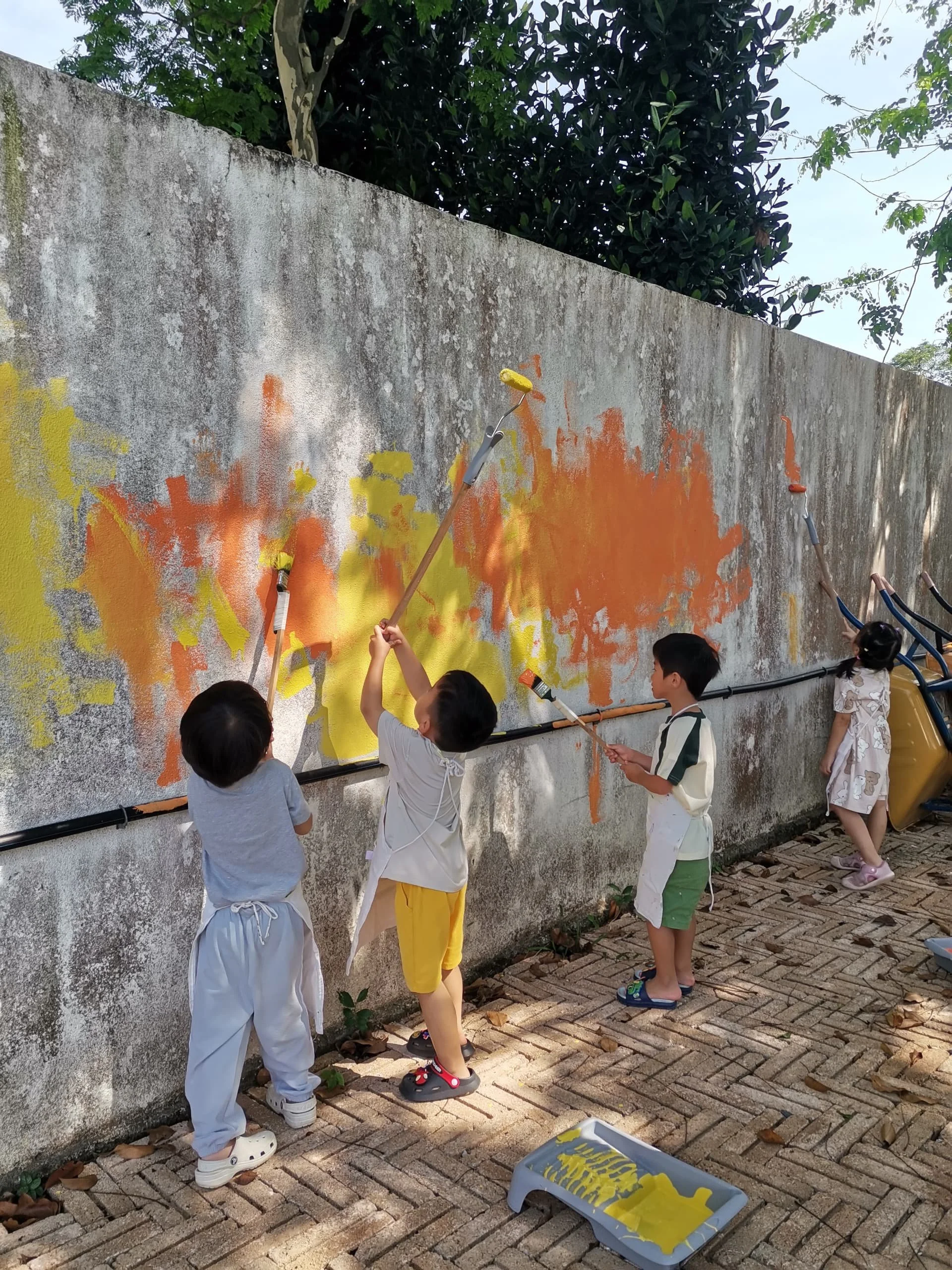 Children painting a large wall with yellow and orange paint outdoors, standing on brick pavement, with trees and a concrete wall in the background.
