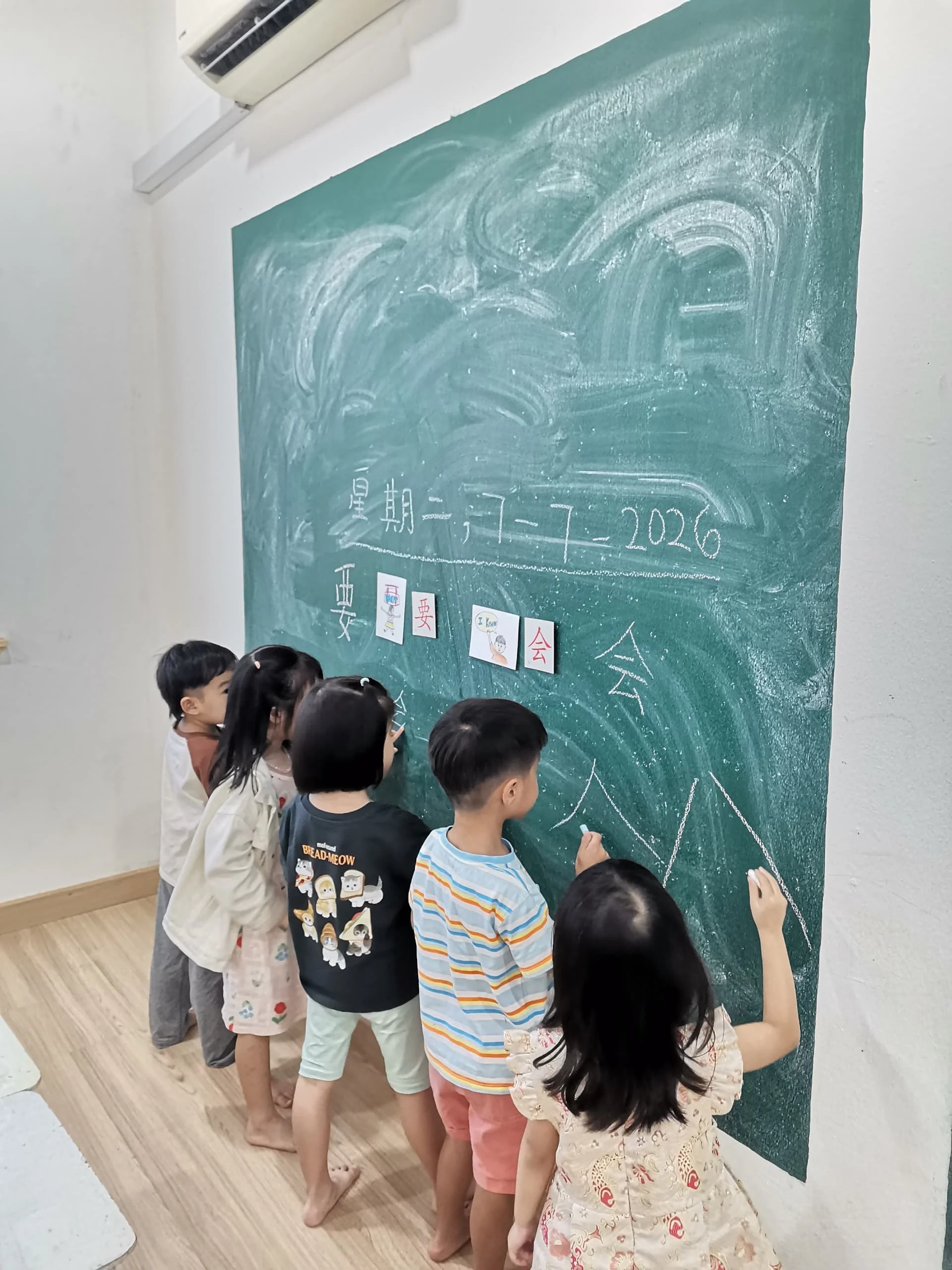 Several children standing in a line, drawing on a green chalkboard. The children are in casual clothing, and the classroom has wooden flooring and a wall-mounted air conditioning unit.