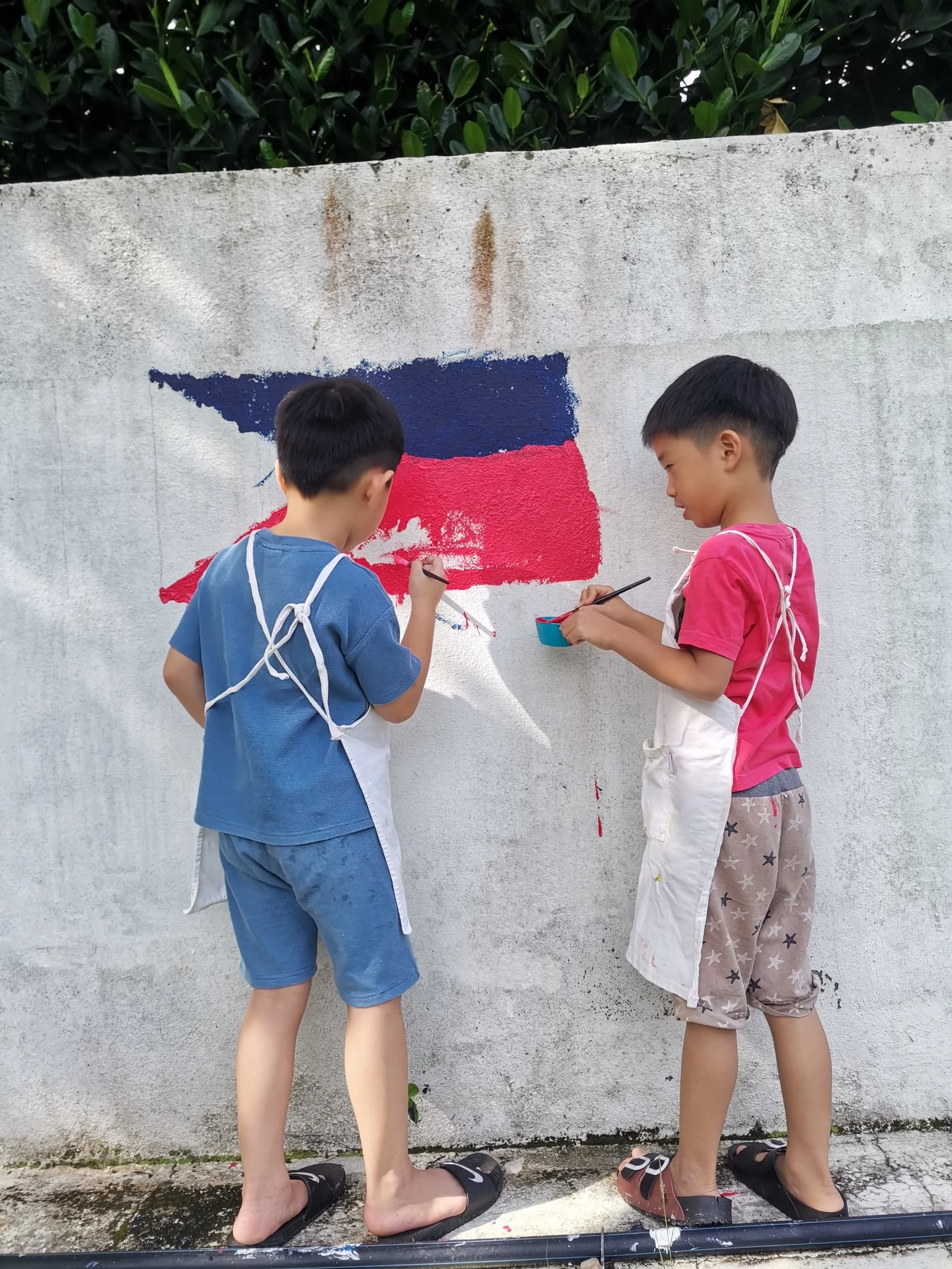 Two children painting a mural on a concrete wall. The mural depicts the flag of Russia with horizontal stripes of white, blue, and red. The children are wearing aprons and holding paintbrushes.