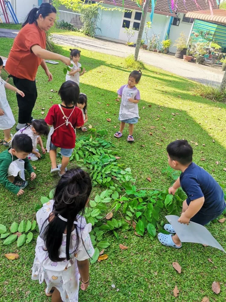 A group of children and a woman outdoors in a grassy area, assembling a long line of green leaves, likely for a craft activity.