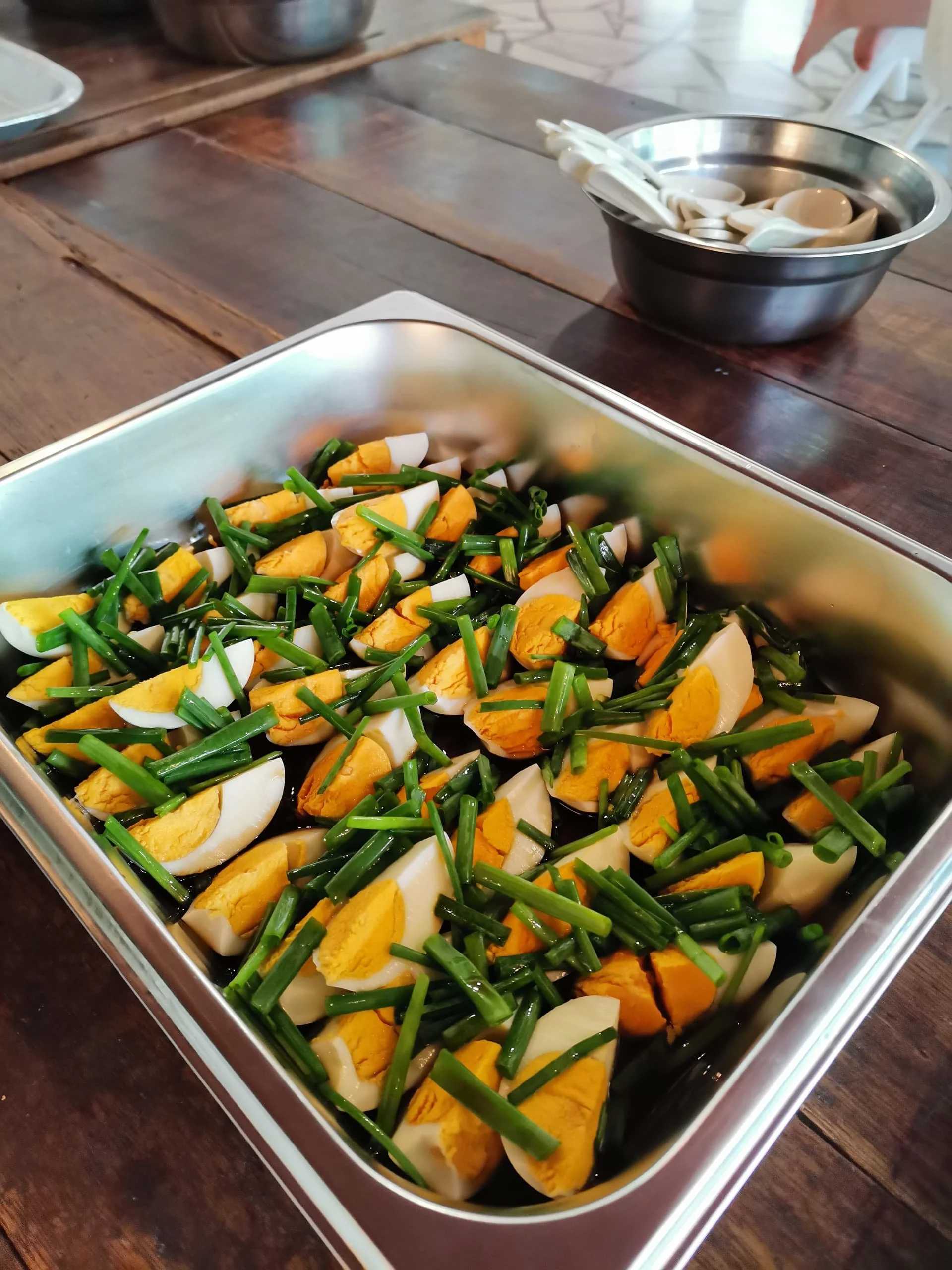 A metal tray filled with chopped hard-boiled eggs, chopped green onions, and possibly a salad mixture. In the background, a stainless steel bowl containing chopped or sliced white vegetables, possibly onions, on a rustic wooden table.