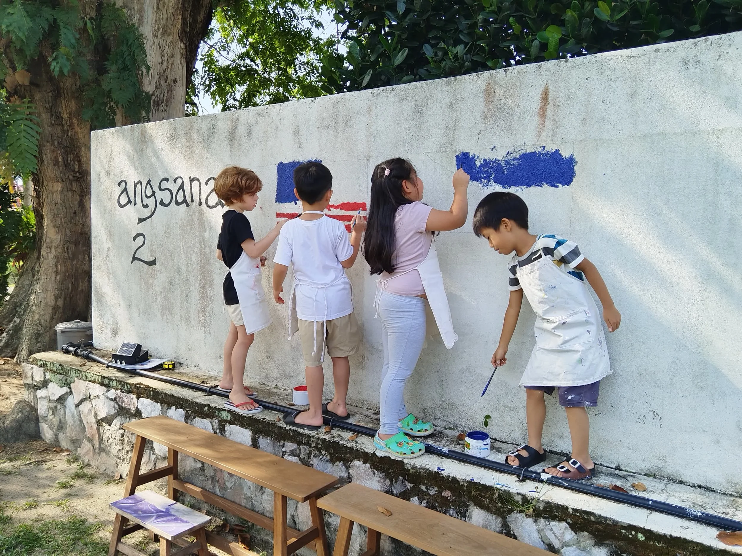 Children painting a mural on a white wall, standing on a narrow ledge outdoors, with trees and greenery in the background.