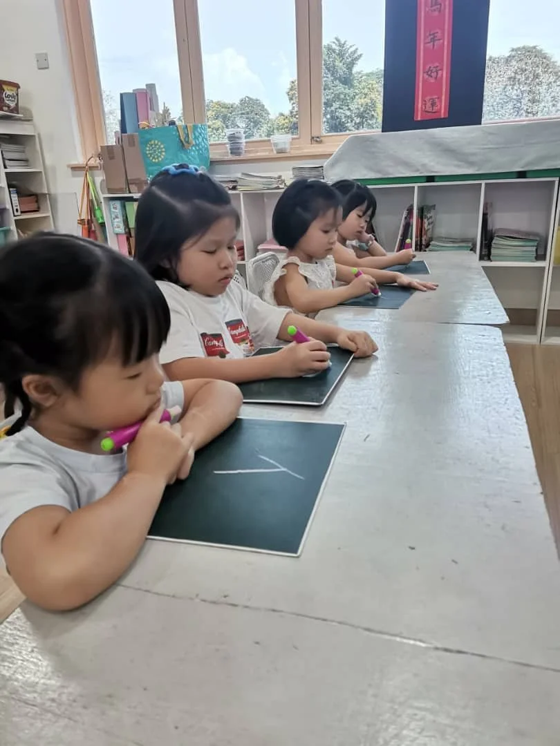 Five young children sitting at a classroom table, each practicing writing on small blackboards with colorful markers, with shelves and windows in the background.