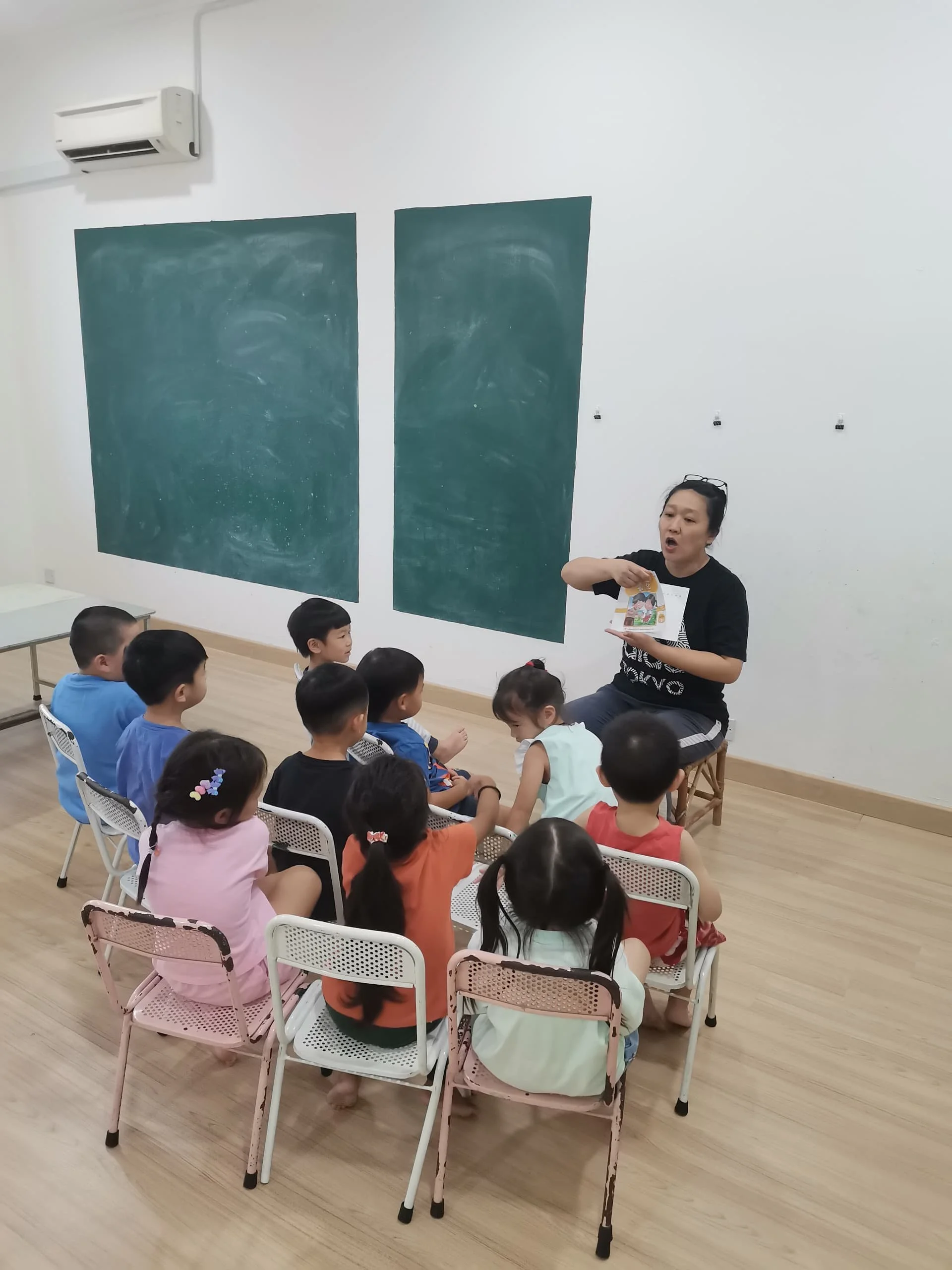A woman is reading a story to a group of children in a classroom. The children are seated on pink and white chairs, facing the woman. The classroom has two large green chalkboards on the wall, and an air conditioner is mounted near the ceiling.