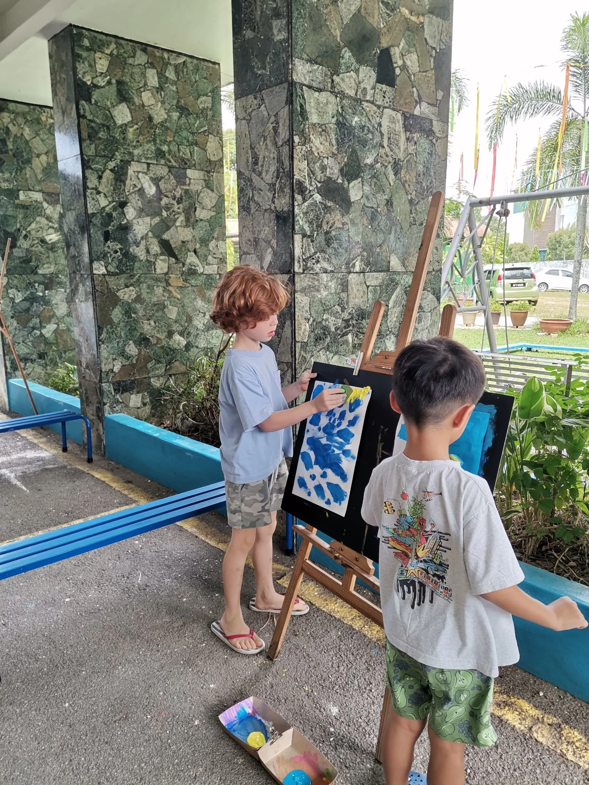 Two children standing outside near a wall decorated with stone tiles, painting on black canvases mounted on wooden easels, with open paint containers on the ground.
