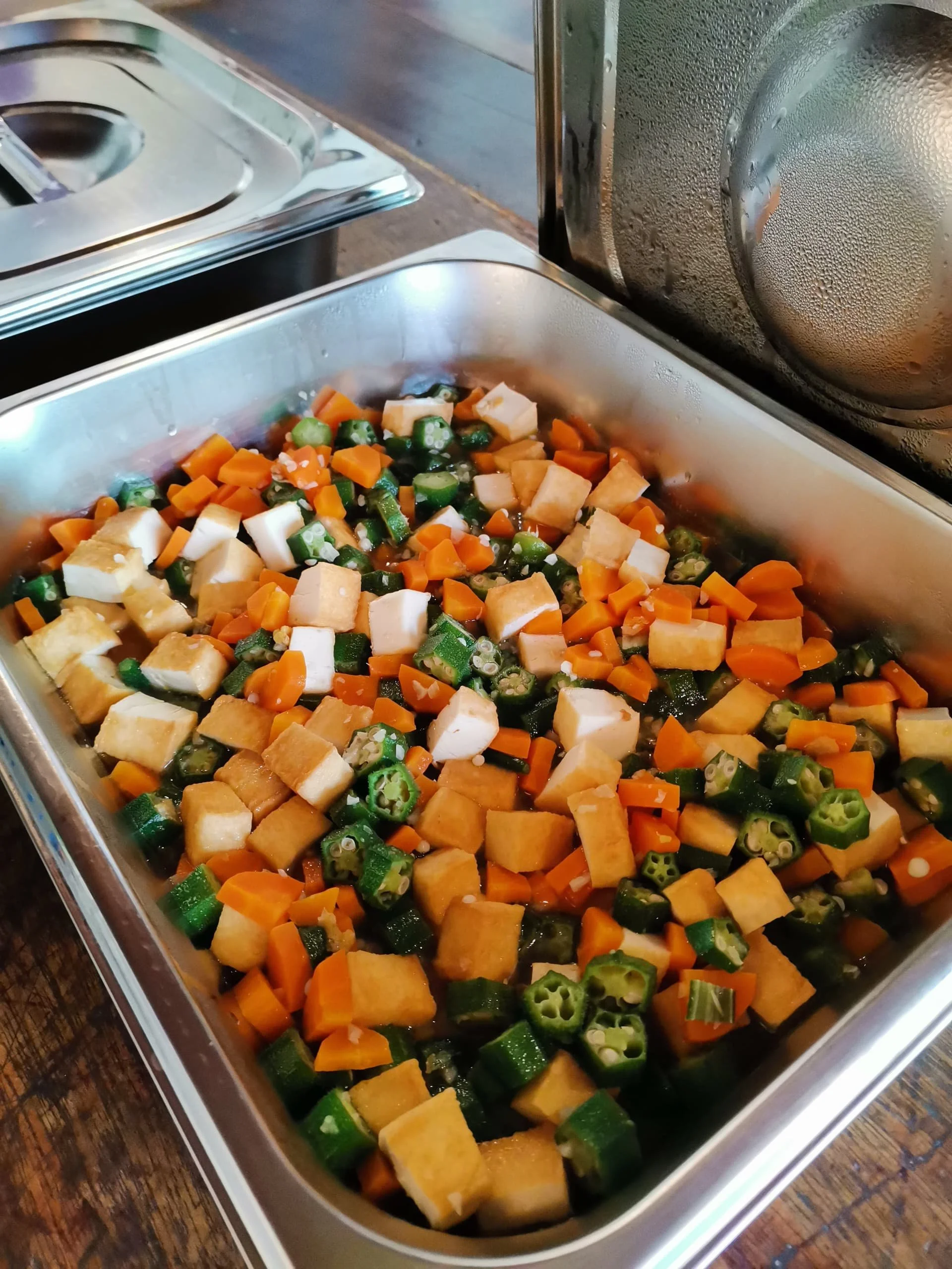 A stainless steel tray filled with chopped vegetables including zucchini, carrots, and possibly other root vegetables, on a wooden surface.