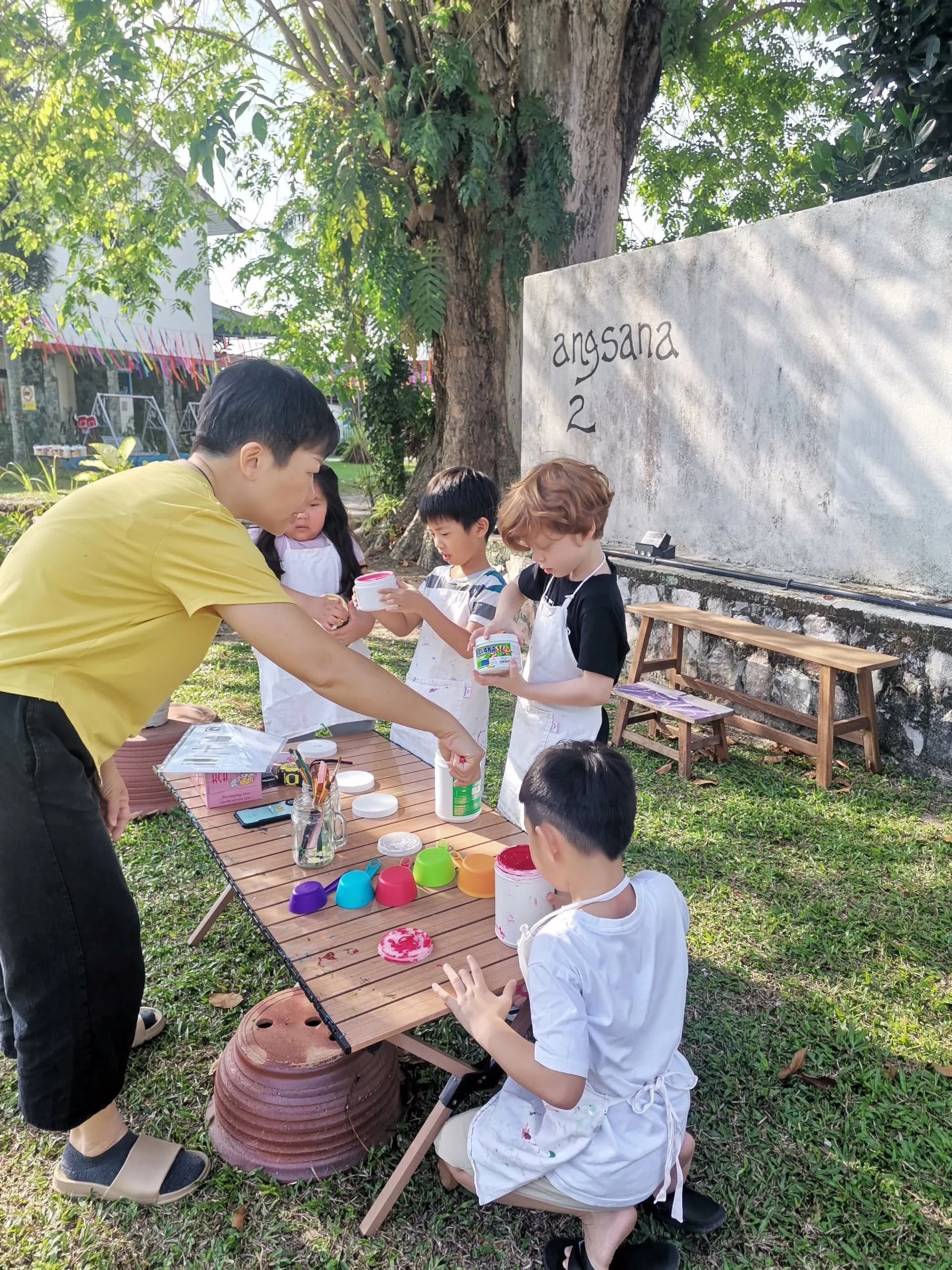 Children and an adult engaged in a craft activity outdoors under a tree, with colorful cups, paint, and art supplies on a wooden table, and a sign that reads 'angsa 2' on a white wall in the background.