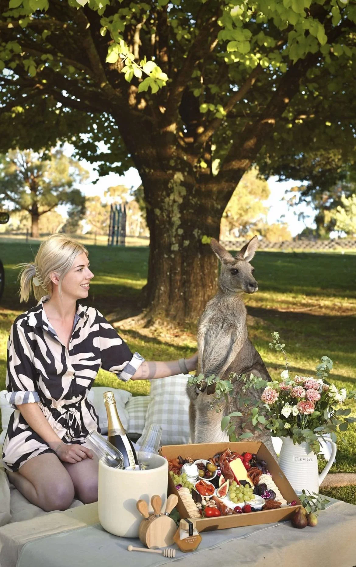 A woman with blonde hair smiling and sitting on a picnic blanket outdoors under a large tree during daytime. She has her right hand on a kangaroo, which is seated beside her. There is a basket of food, a bottle of champagne in an ice bucket, flowers in a pitcher, and various picnic items on the blanket.