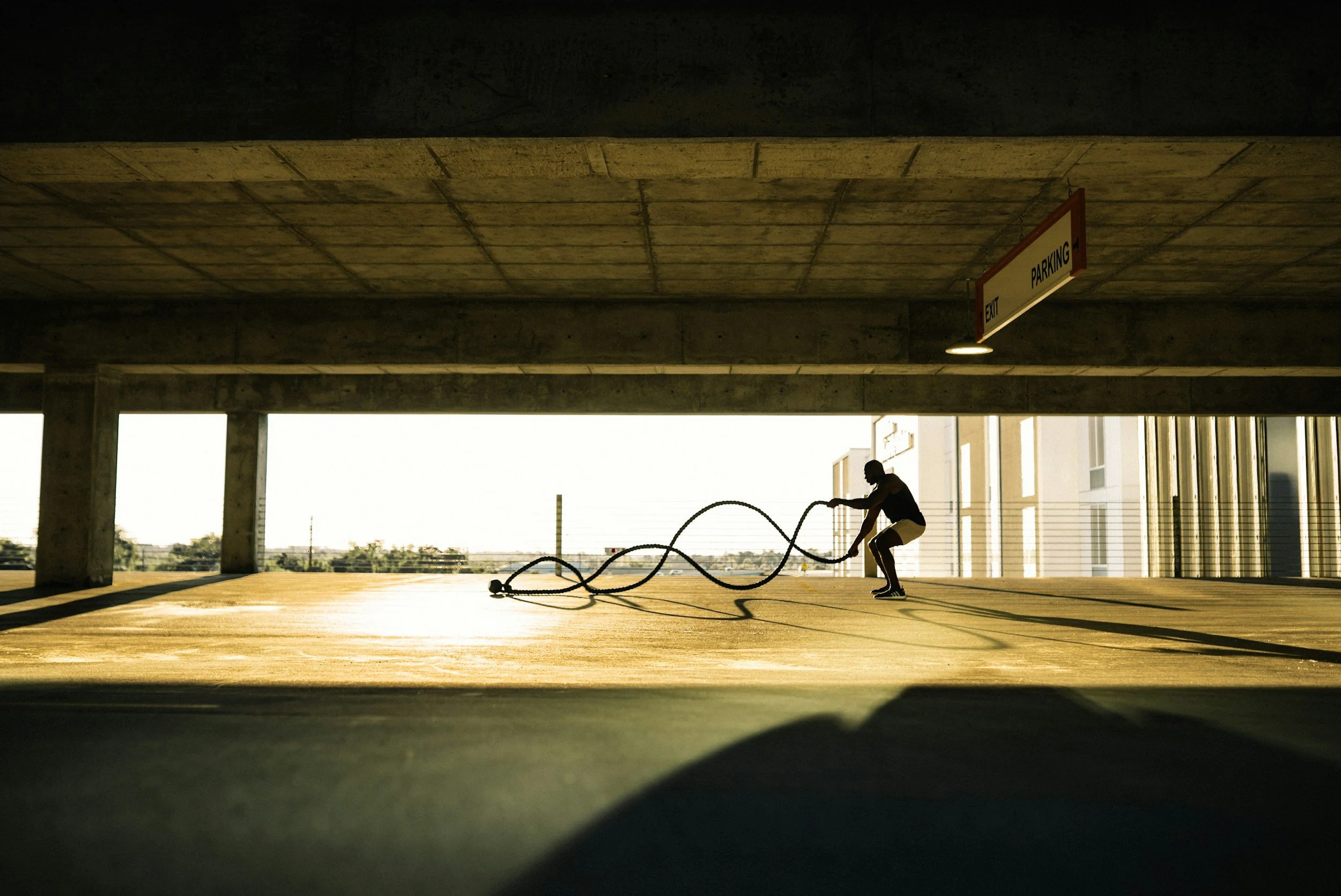 Person exercising with battle ropes in a parking garage, with dramatic lighting and an exit sign overhead.