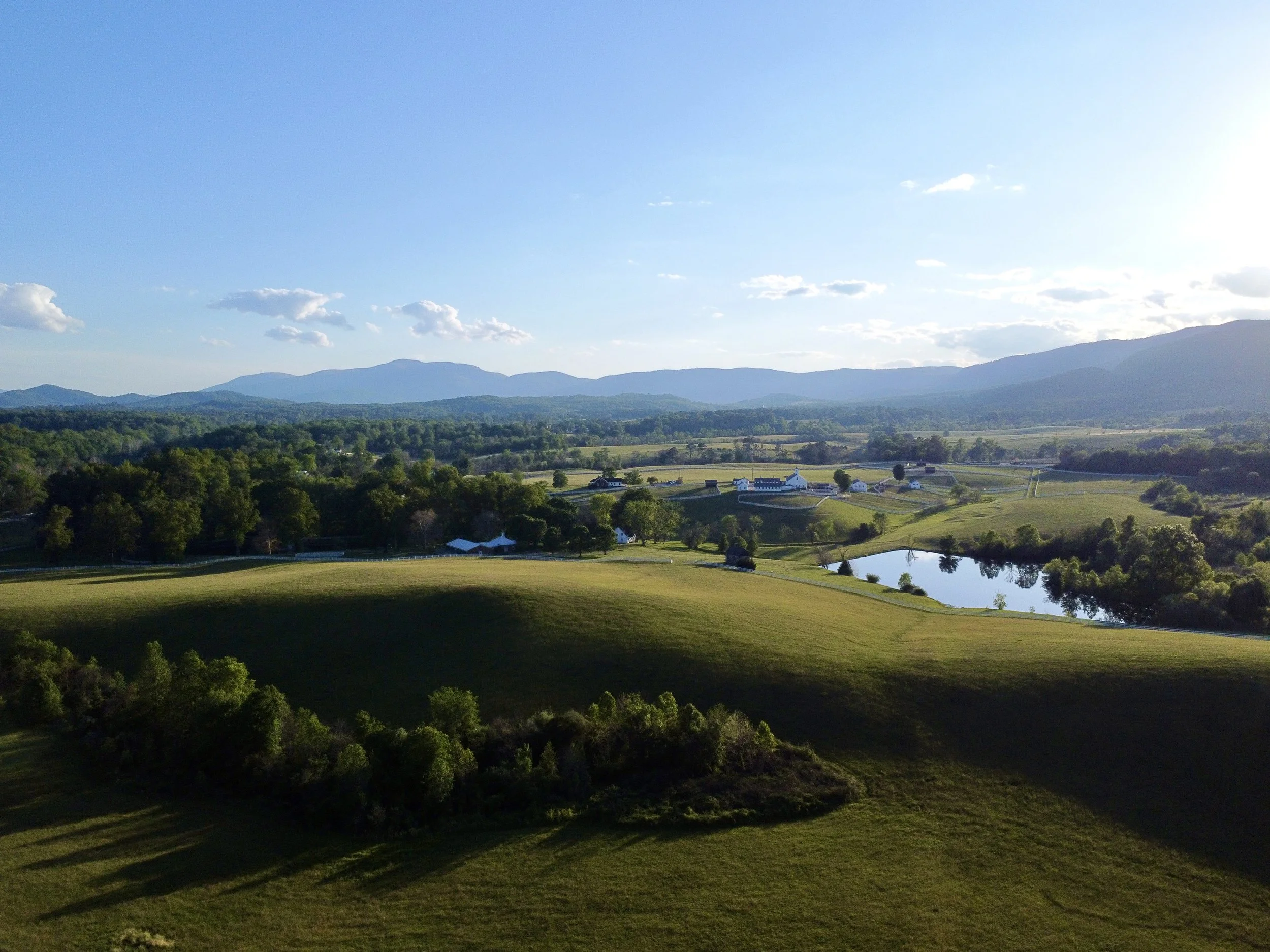 A scenic landscape of rolling green hills, with a few white farm buildings and a pond, set against distant blue mountains under a partly cloudy sky.