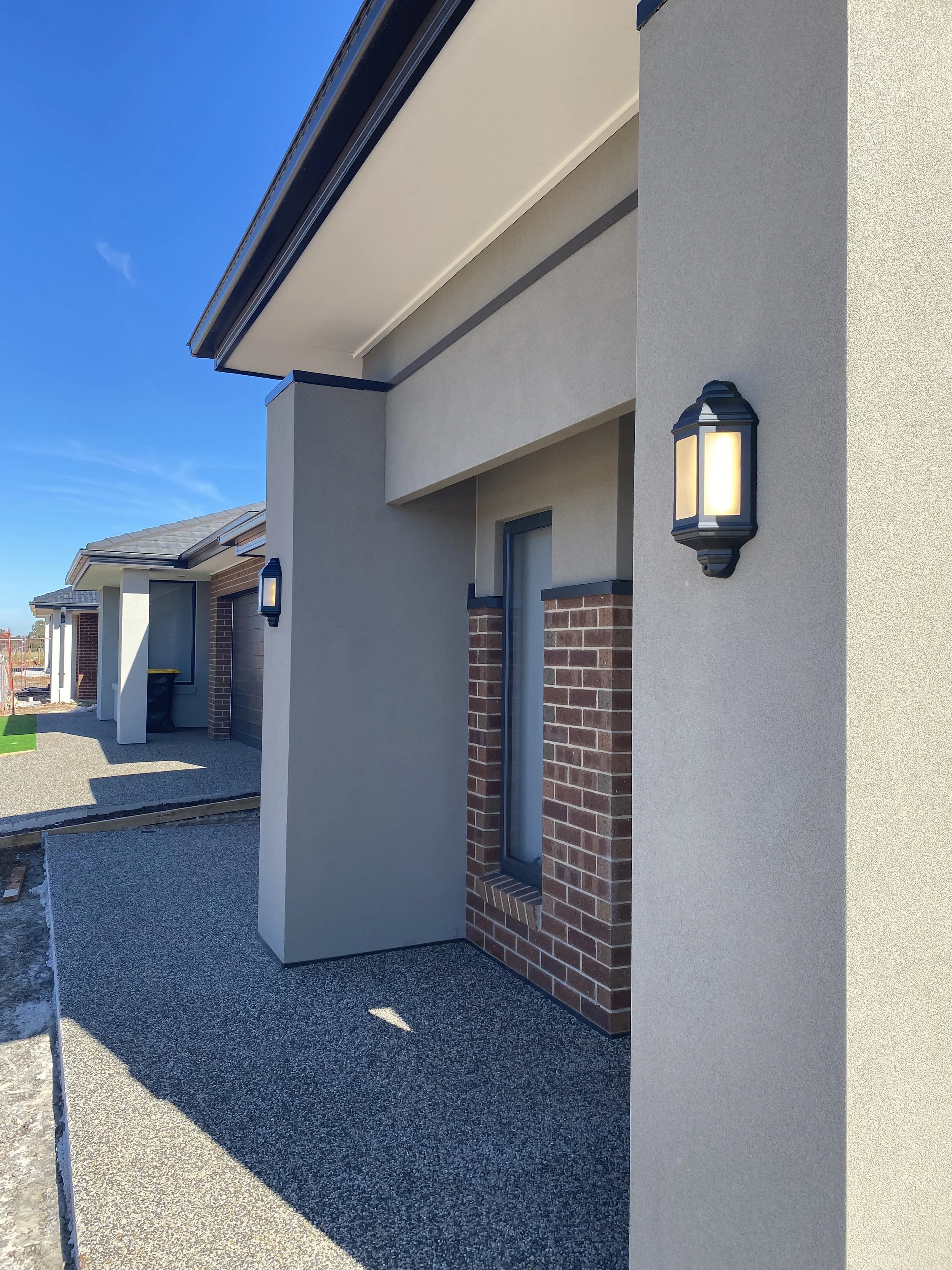 Exterior view of a modern house with wall-mounted outdoor lights, brick accents, and a textured concrete porch under a clear blue sky.