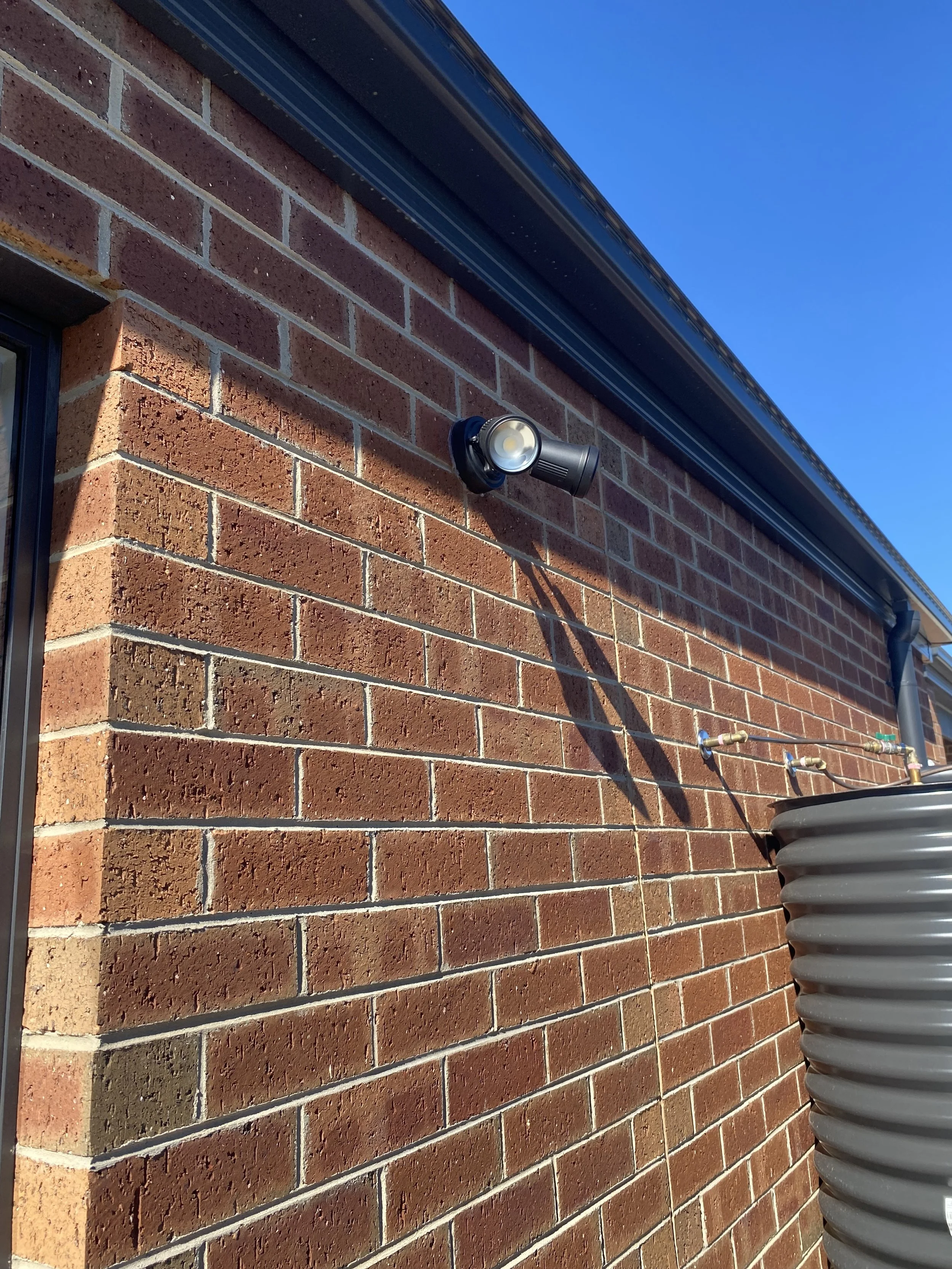 Exterior brick wall of a building with a security camera and water piping, under a blue sky.