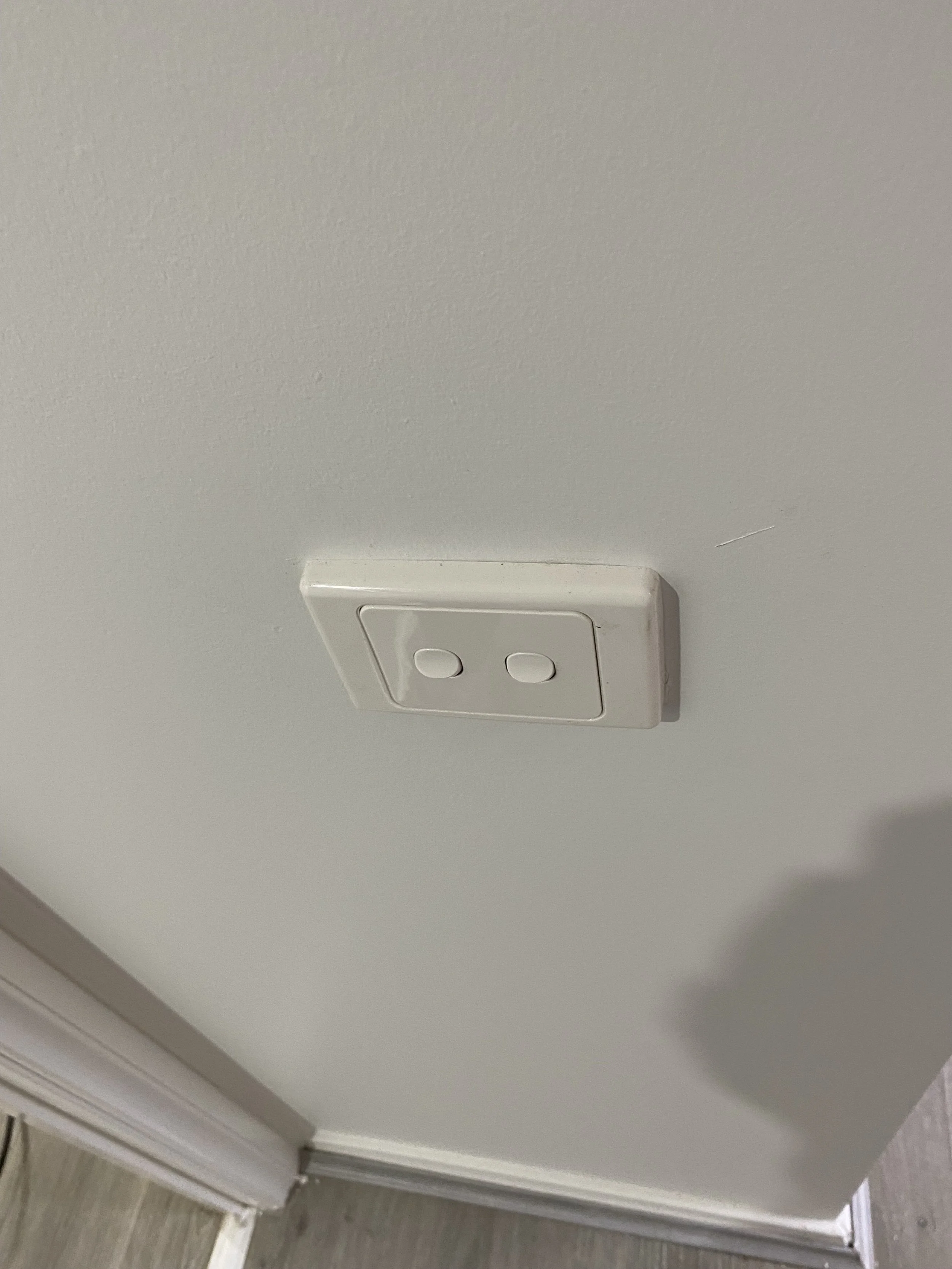 White electrical outlet with two circular holes on a plain ceiling, with a shadow cast on the ceiling and a corner of a wall and floor visible.