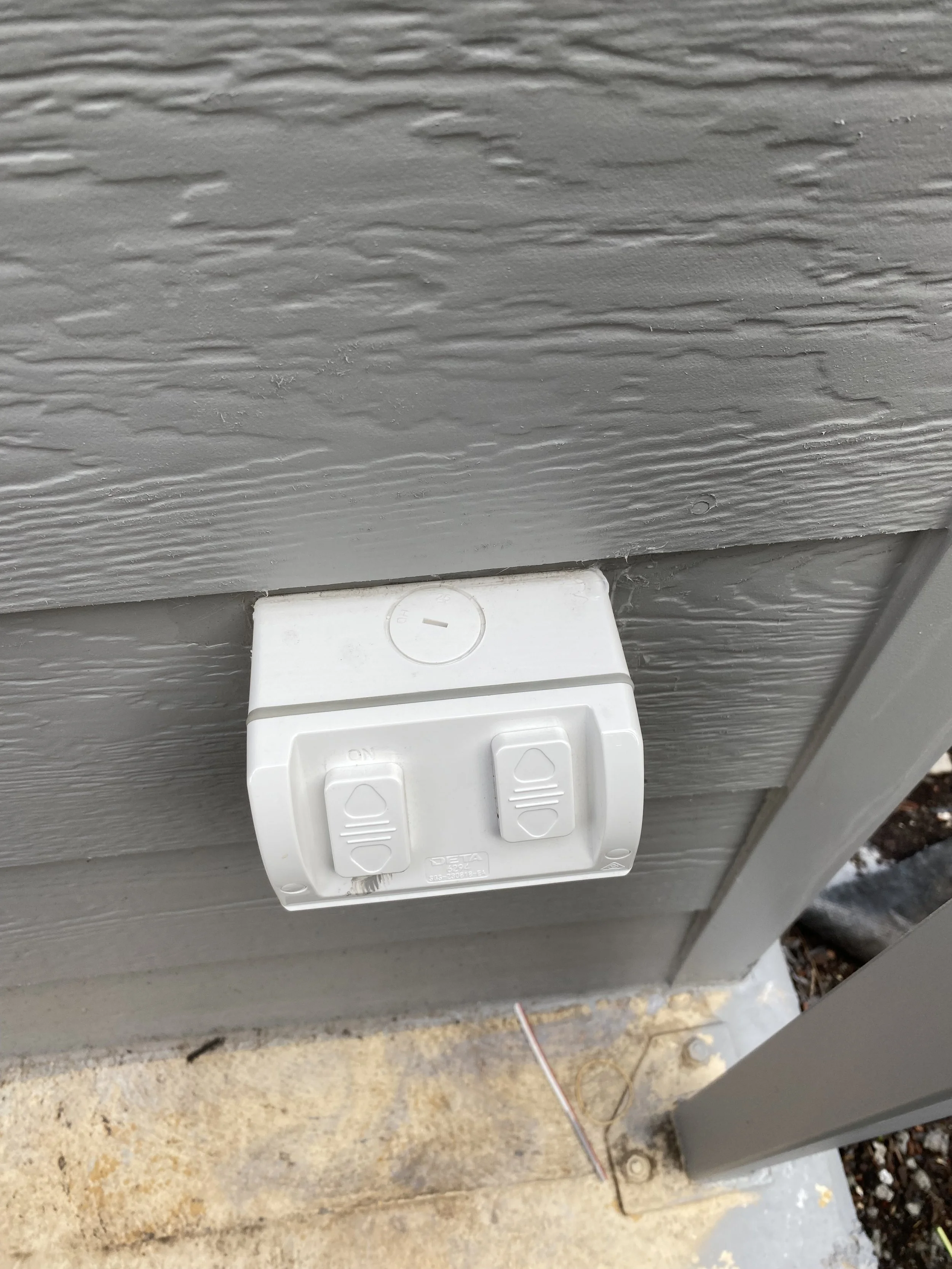 An outdoor electrical outlet with two switches mounted on a gray wooden wall and a concrete surface below.