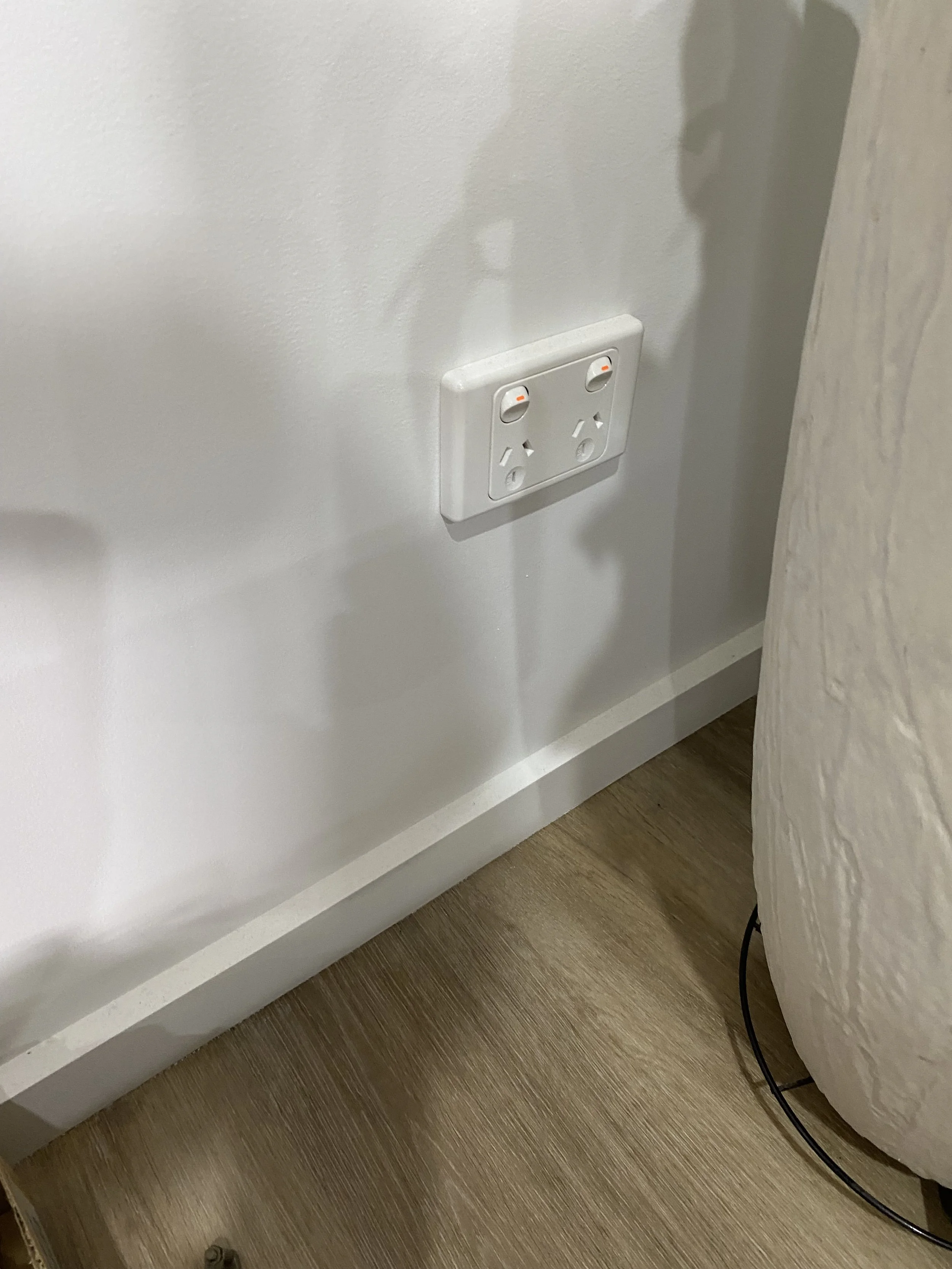 Electrical outlet with two switches and three sockets on a white wall near a bed and wooden floor.
