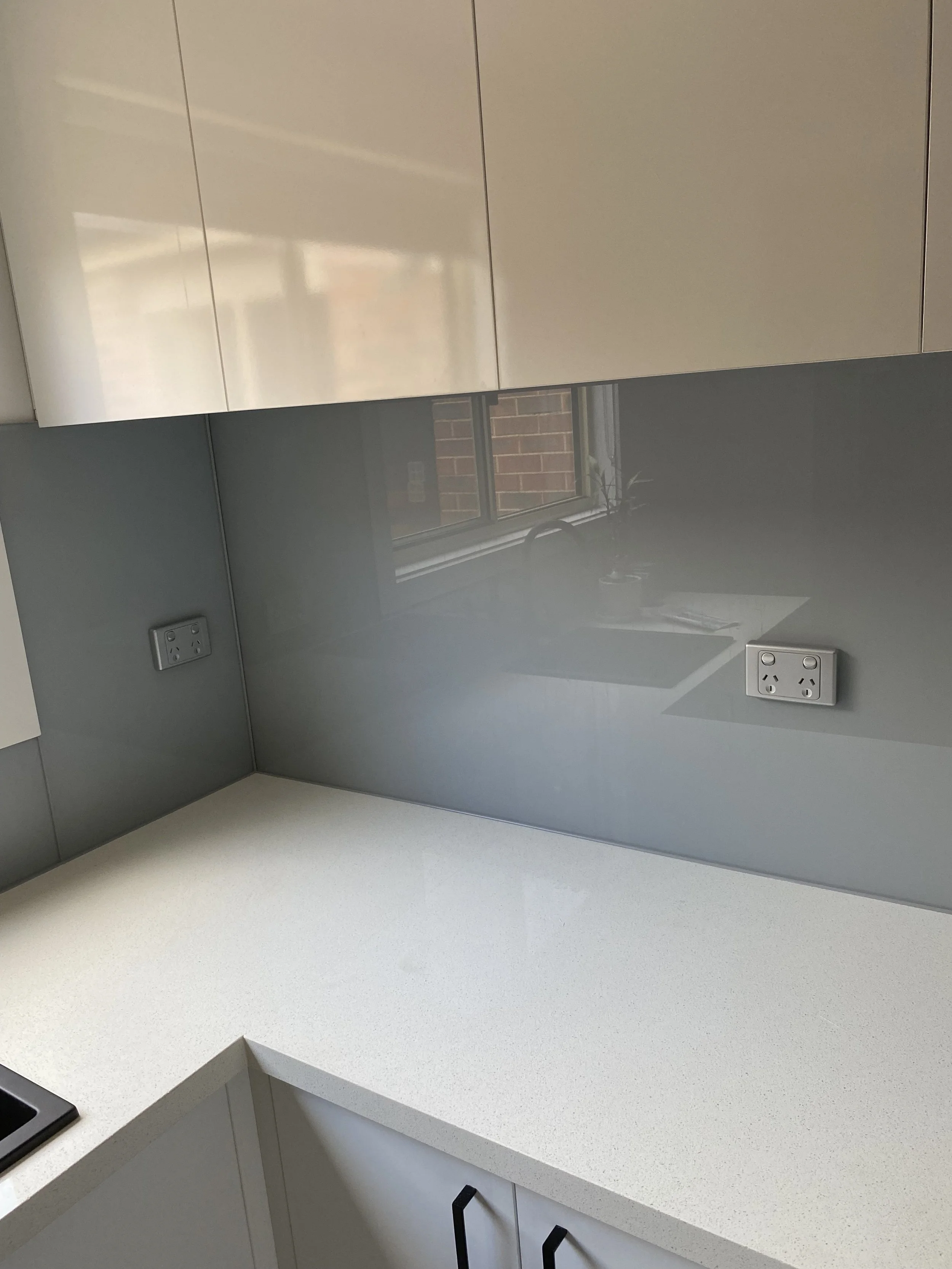 Kitchen with white countertop, beige cabinets, gray reflective backsplash, and two two-plug electrical outlets.
