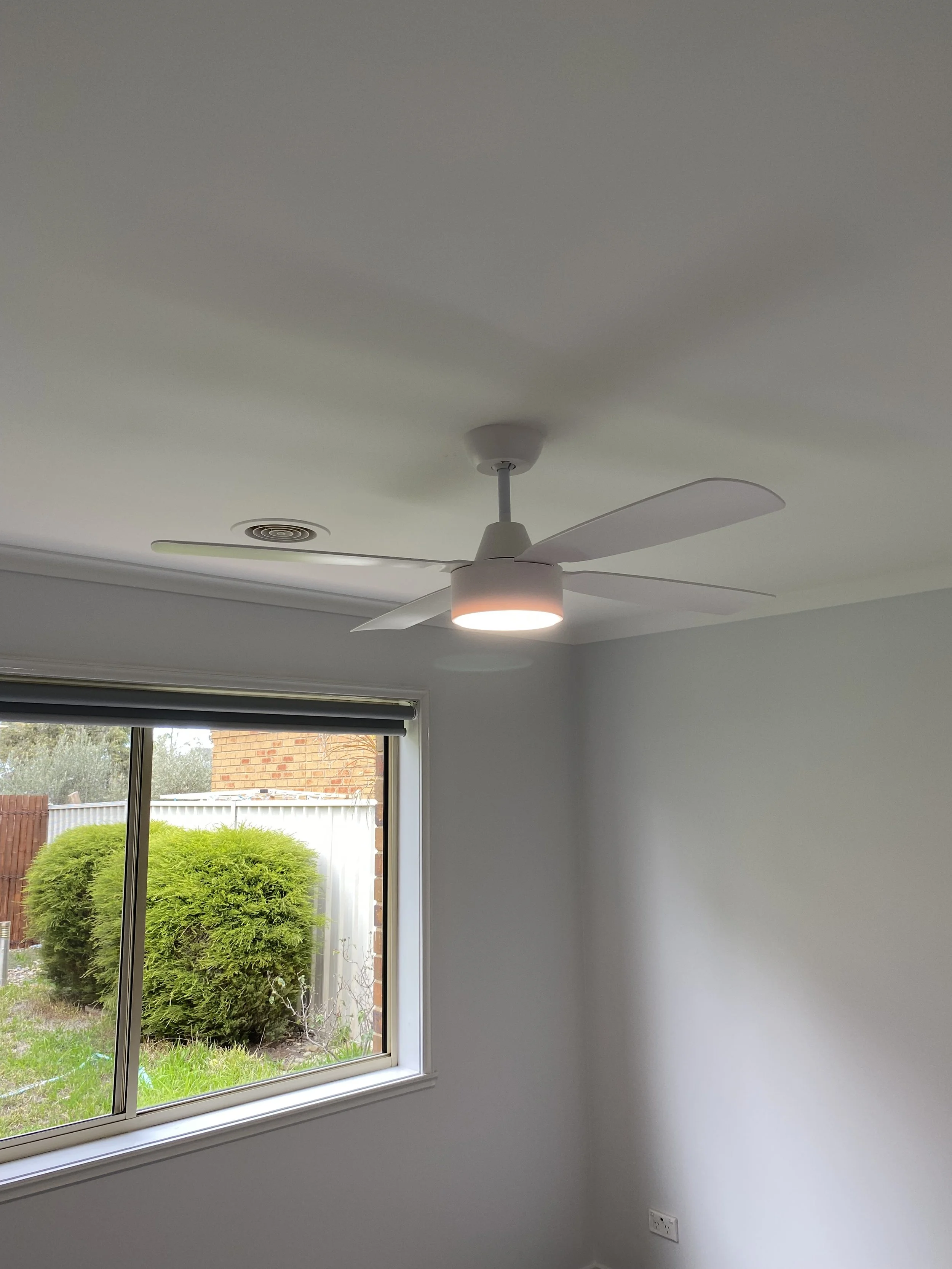 A white ceiling fan with a built-in light in a room with white walls and a window showing a bush and a brick wall outside.