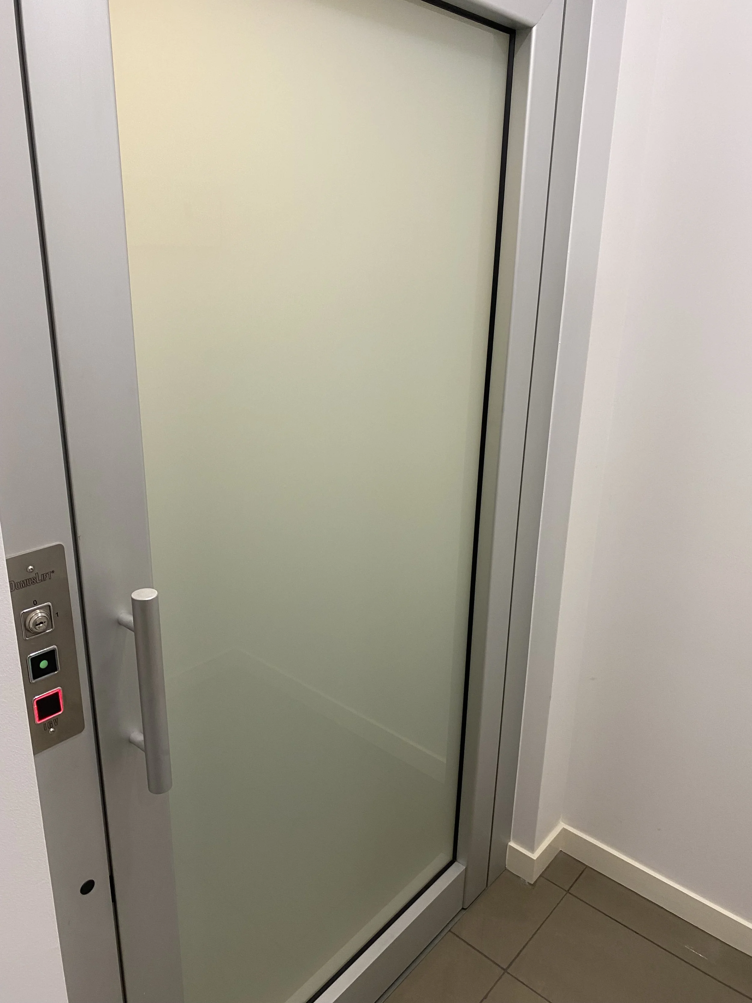 A glass elevator door with a silver handle, control buttons, and a frosted glass panel. The wall and floor surrounding the elevator are white and beige respectively.