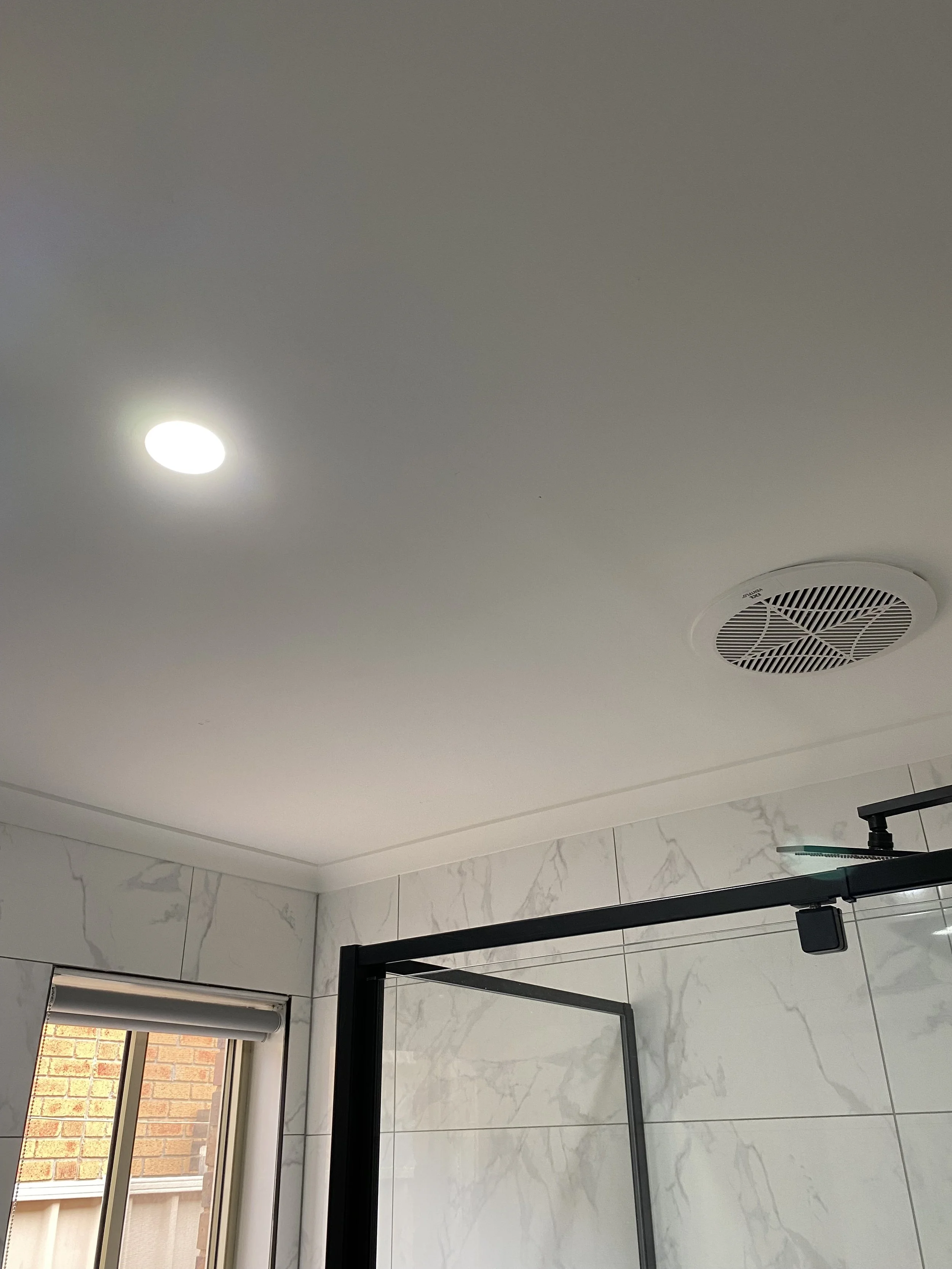 Ceiling of a bathroom with a recessed light, circular exhaust vent, part of a shower door, window, and marble tiles on the wall.
