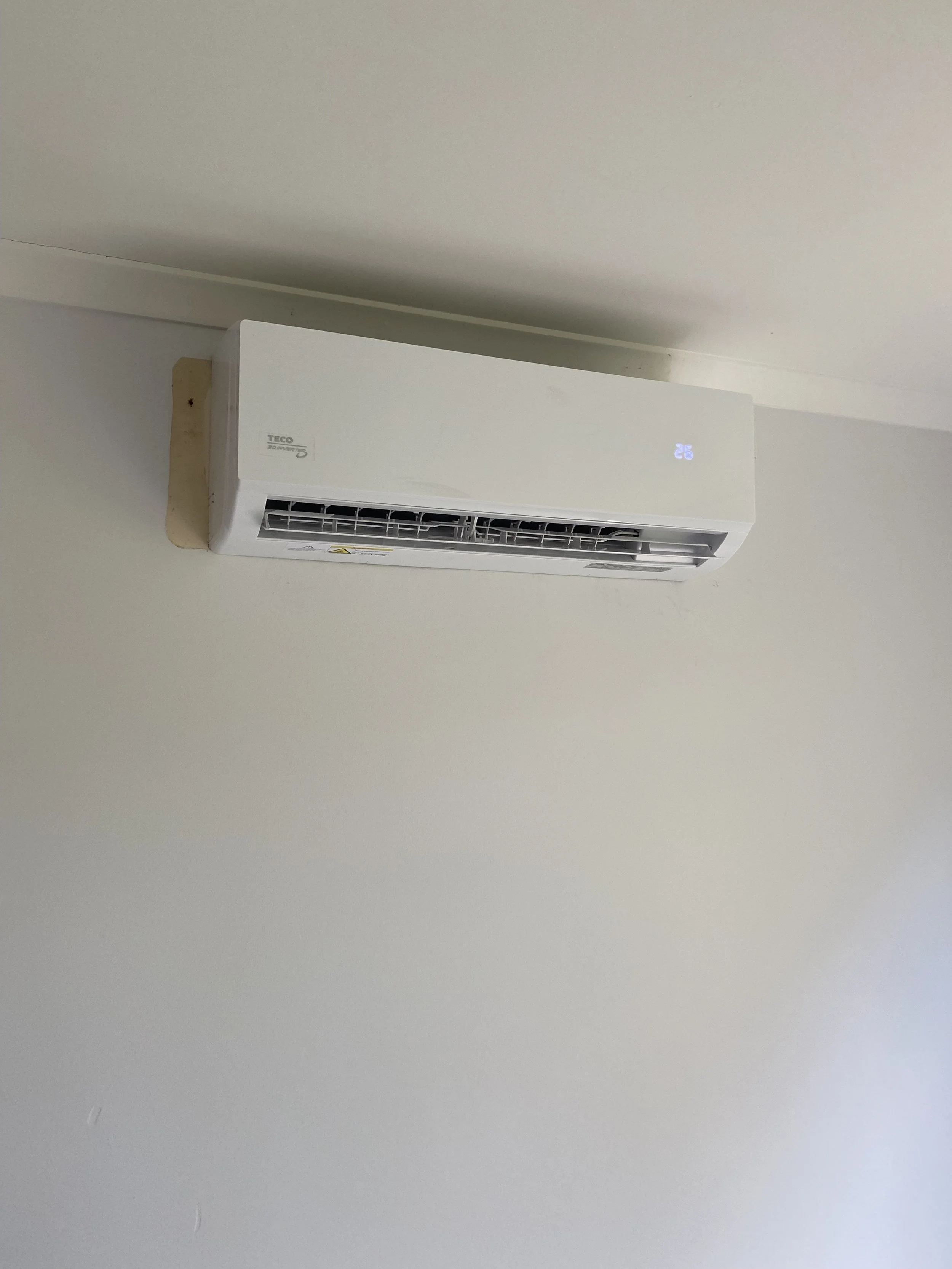 Wall-mounted white air conditioning unit with a digital display showing the temperature set to 26 degrees, installed on a cream-colored wall near the ceiling.