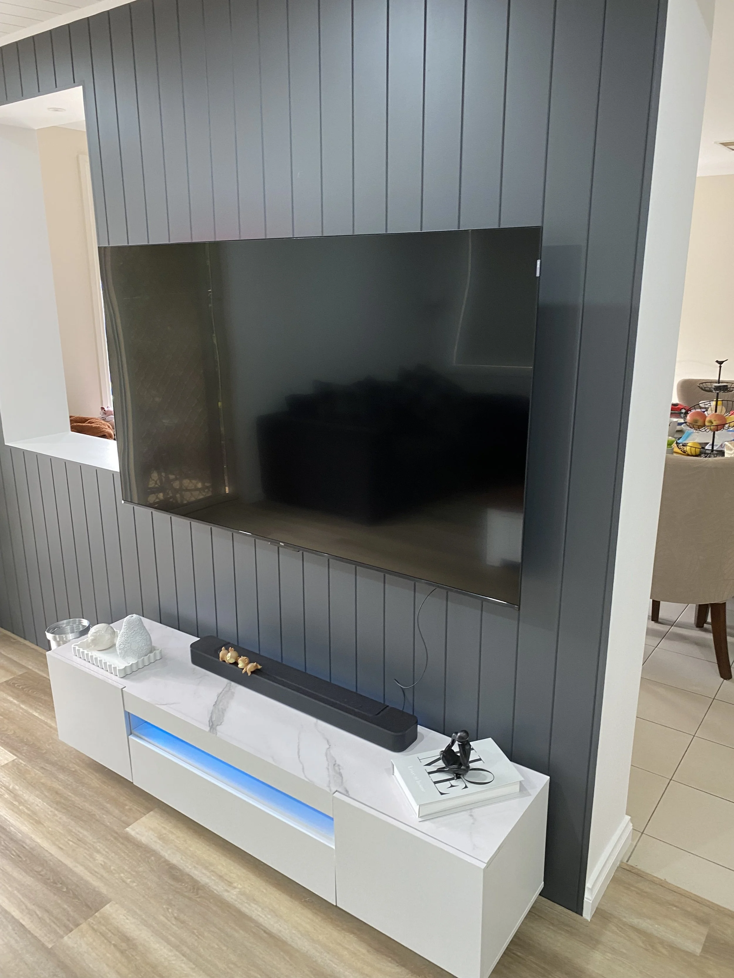 Living room entertainment area with a wall-mounted flat-screen TV, a white console table with decorative items, and a bluish led light strip underneath.