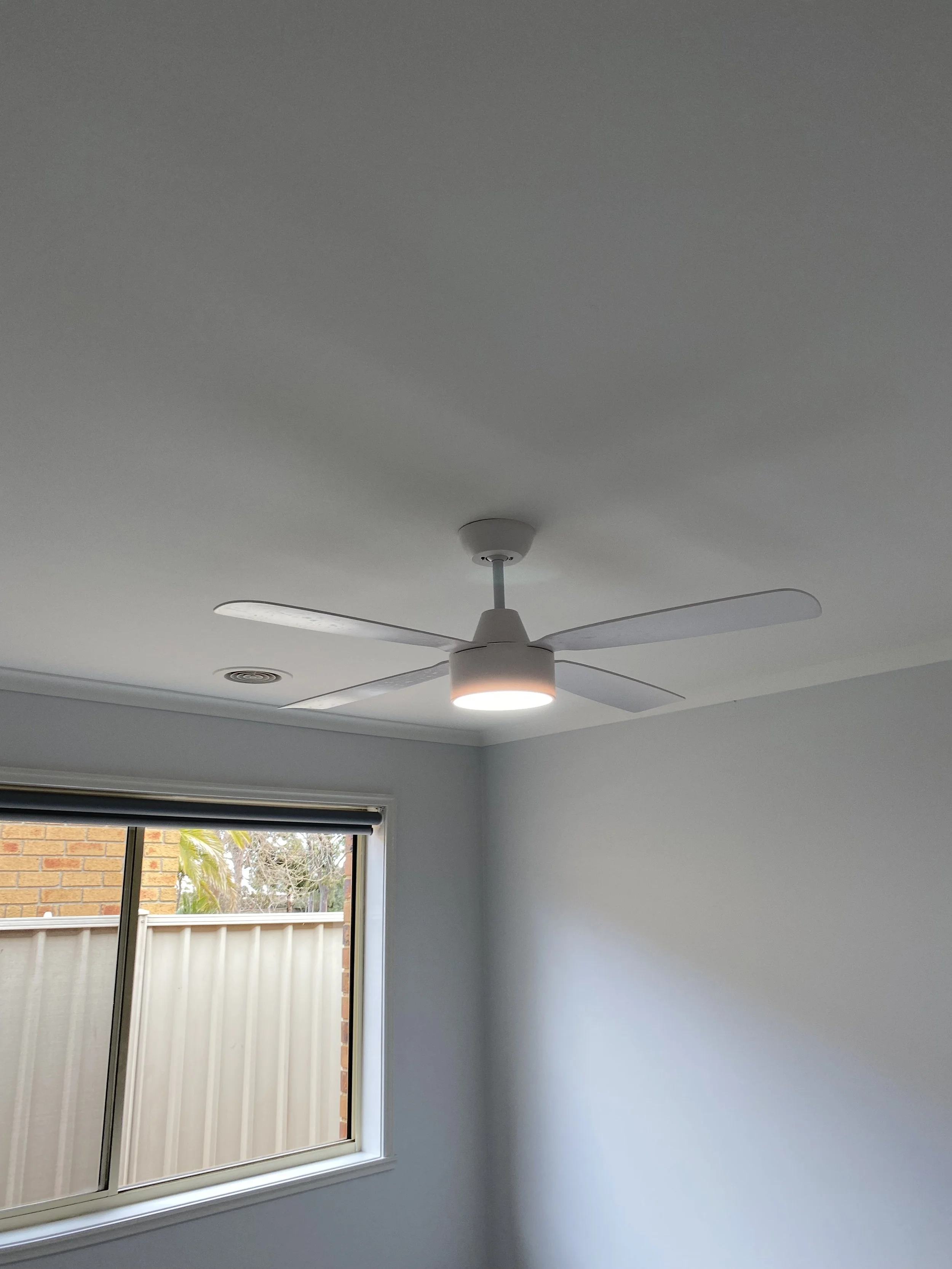 White ceiling fan with lights mounted on a white ceiling, next to a window with a view of a brick wall, fence, and outdoor trees.