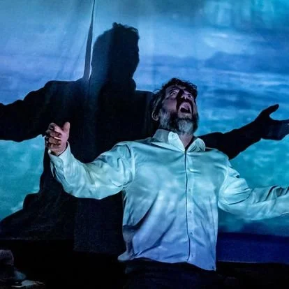A man with a beard, wearing a white shirt, standing with arms open and looking upwards, with a large shadow of a person projected behind him. Gold Coast Theatre Thursday's Child