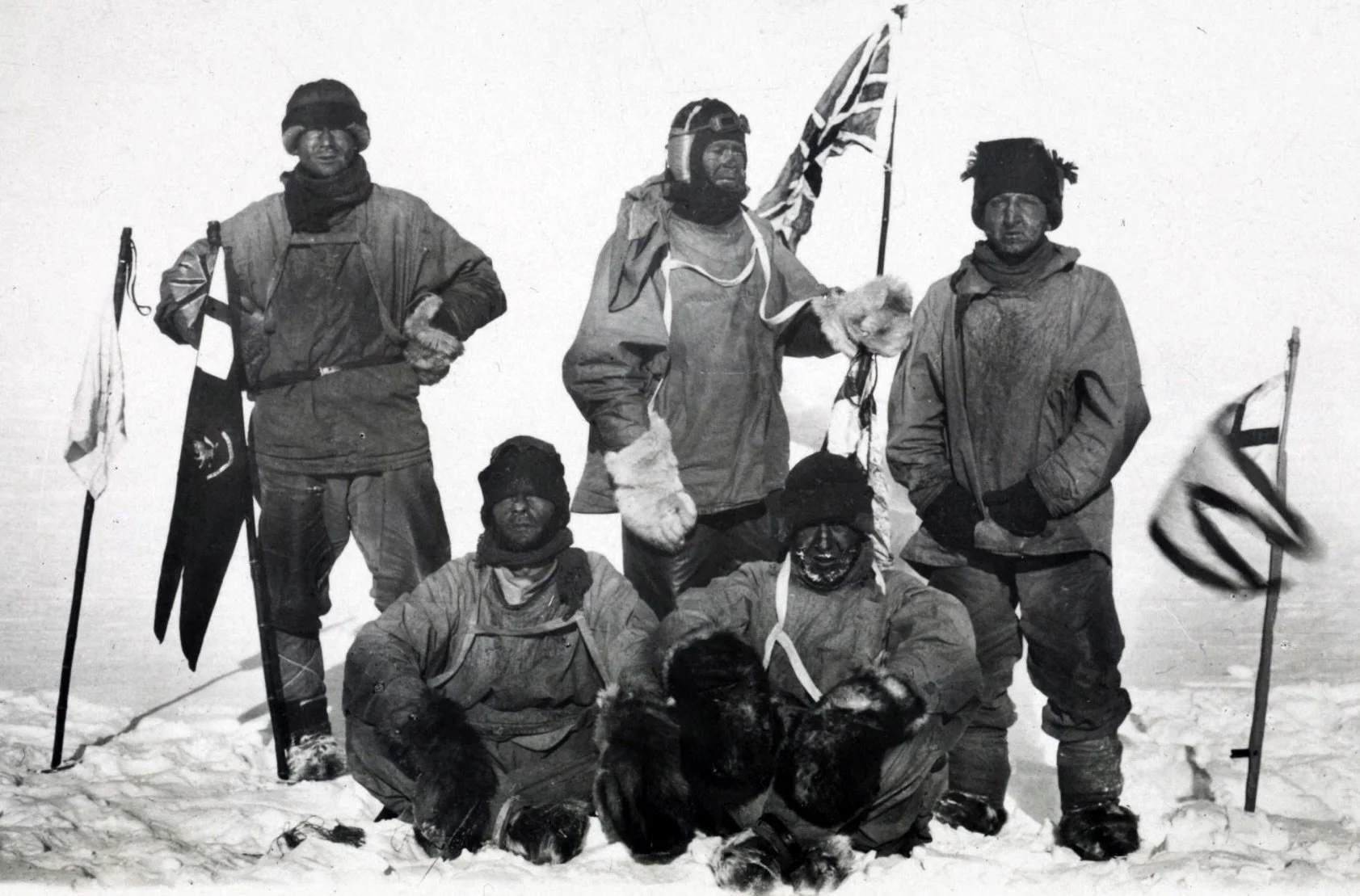 Black and white photo of six explorers dressed in heavy winter gear, with some holding flags, standing and sitting in snow, suggesting exploration of polar or icy region.