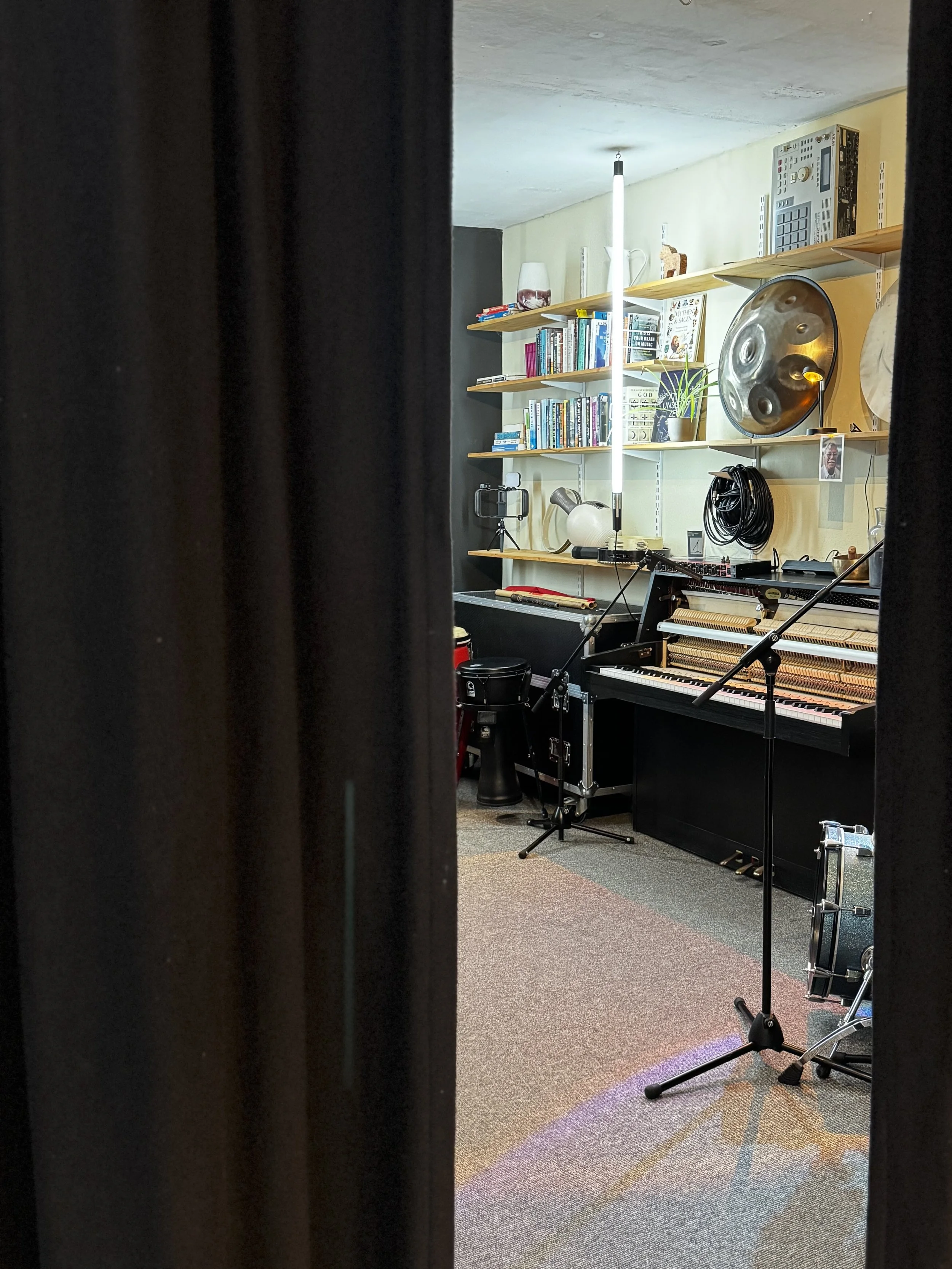 Ein Blick in ein Musikzimmer, das einen Flügel, ein Mikrofonständer, ein Schlagzeug, Regale mit Büchern, Musikinstrumenten und technischen Geräten enthält.