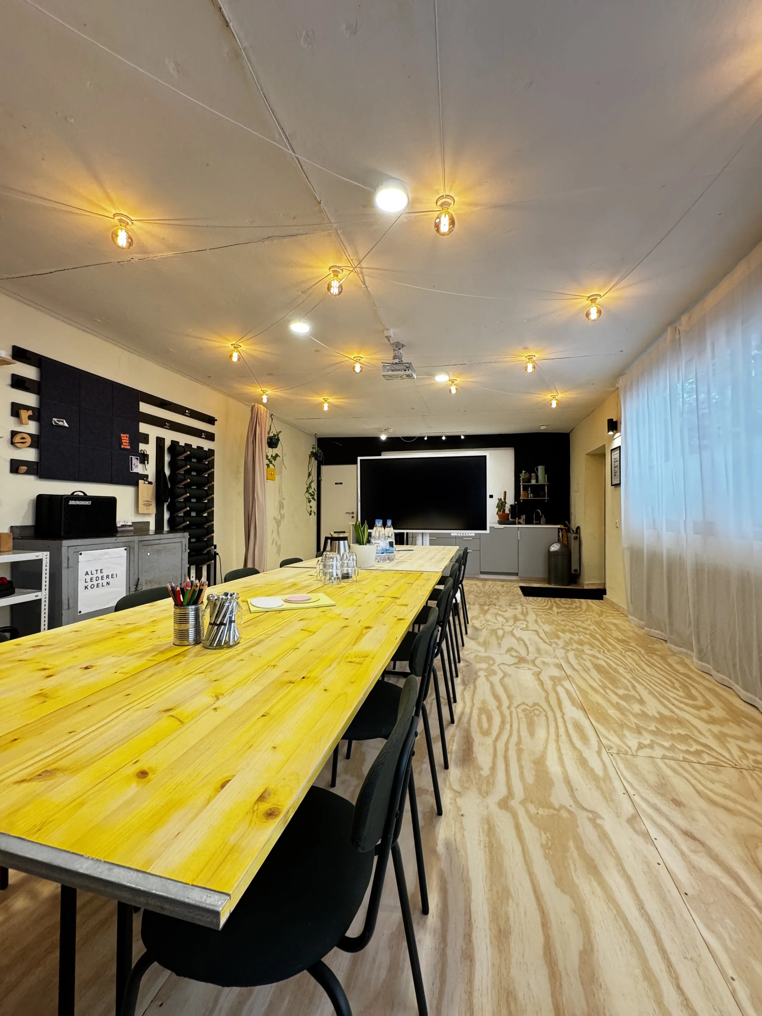 Ein moderner, gut beleuchteter Konferenzraum mit einem langen gelben Tisch, mehreren schwarzen Stühlen, einer großen schwarzen Bildschirme und einer Küchenzeile im Hintergrund. Die Decke ist mit mehreren Leuchtkörpern versehen.