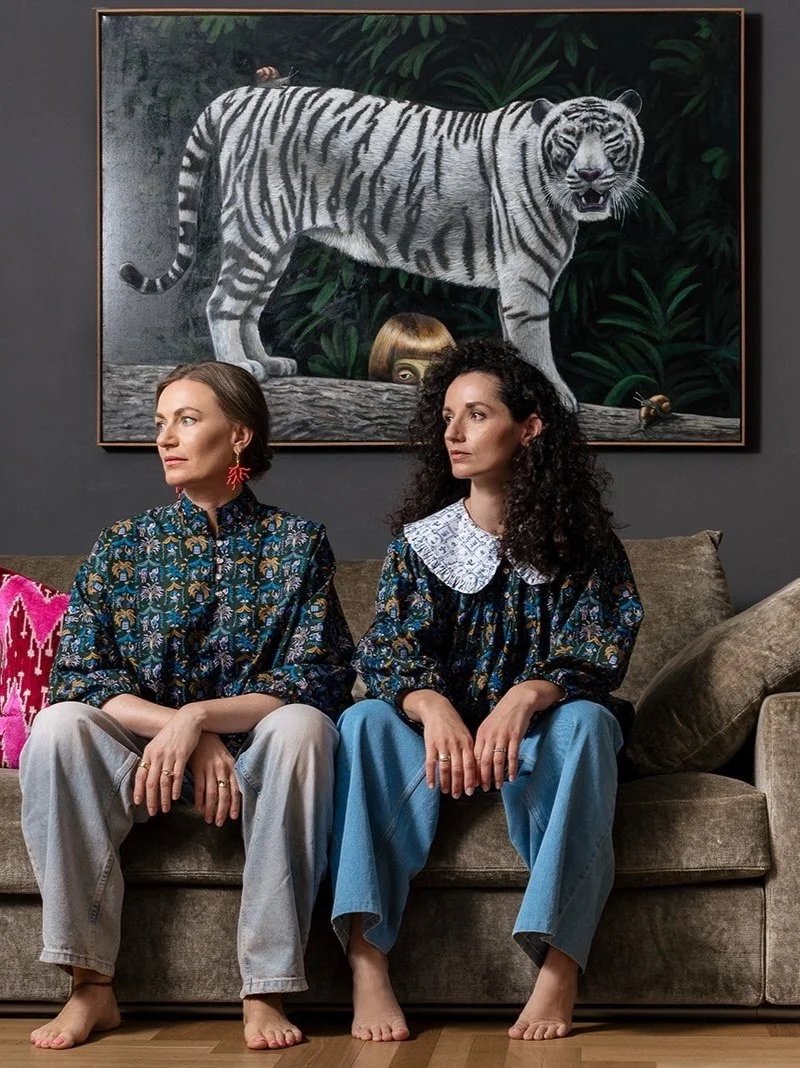 Two women sitting on a beige couch in a living room, with a large painting of a white tiger hanging on a dark gray wall behind them. A tall vase with colorful flowers is on the right side of the couch.