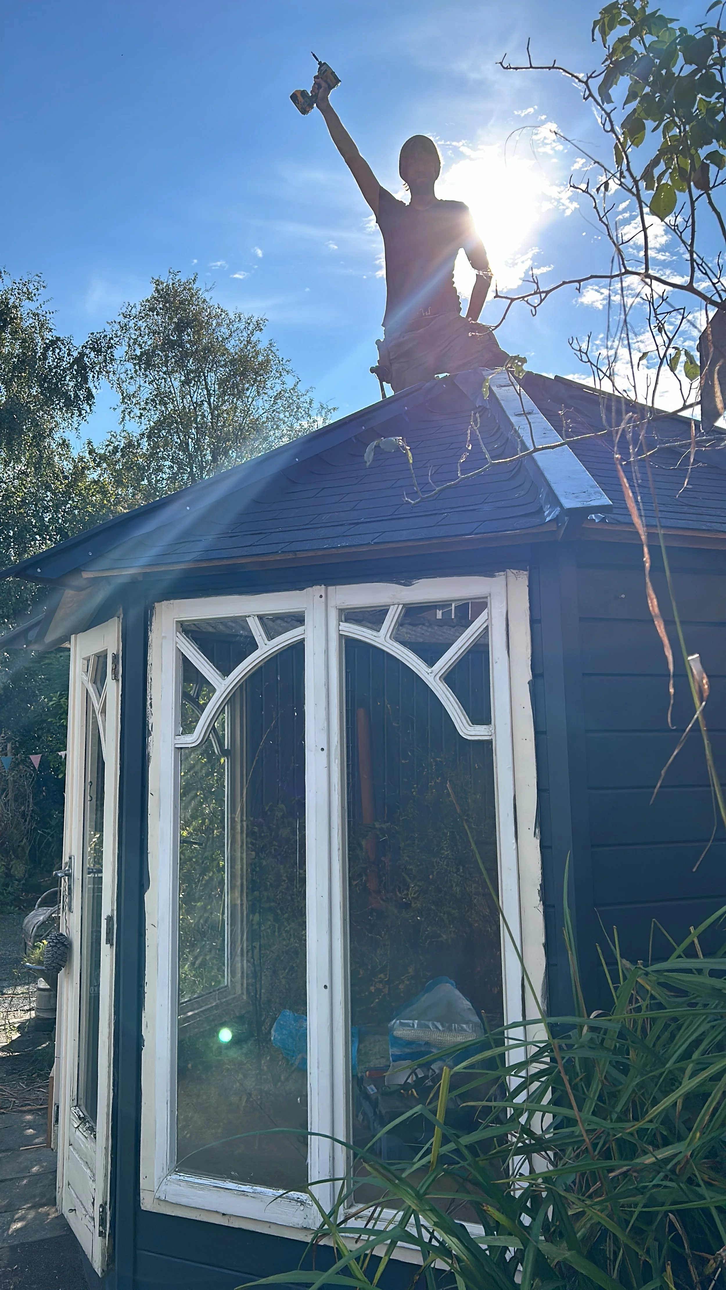 A person standing on the roof of a small house, holding a power drill in the air, with the sun shining behind them and trees in the background.