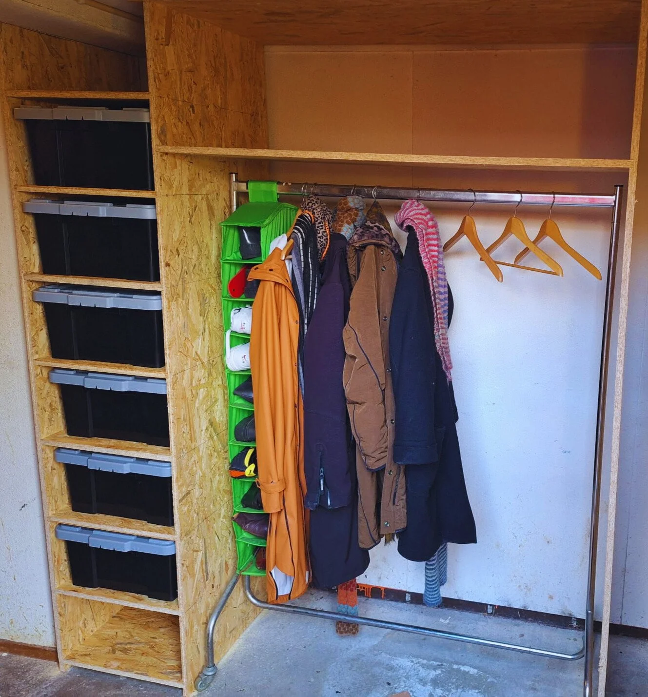 A wooden closet with black storage bins on the left and a clothing rack with jackets and sweaters on the right.