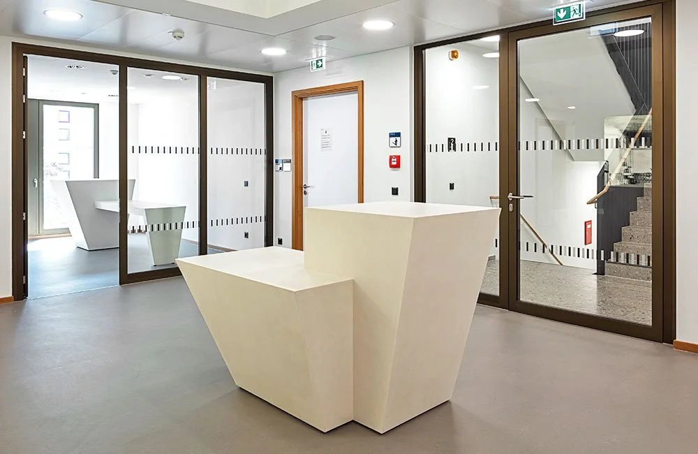 Moderne Innenansicht eines Büros mit einem weißen, geometrischen Empfangstresen aus Designbeton, Glastüren und Treppe im Hintergrund.