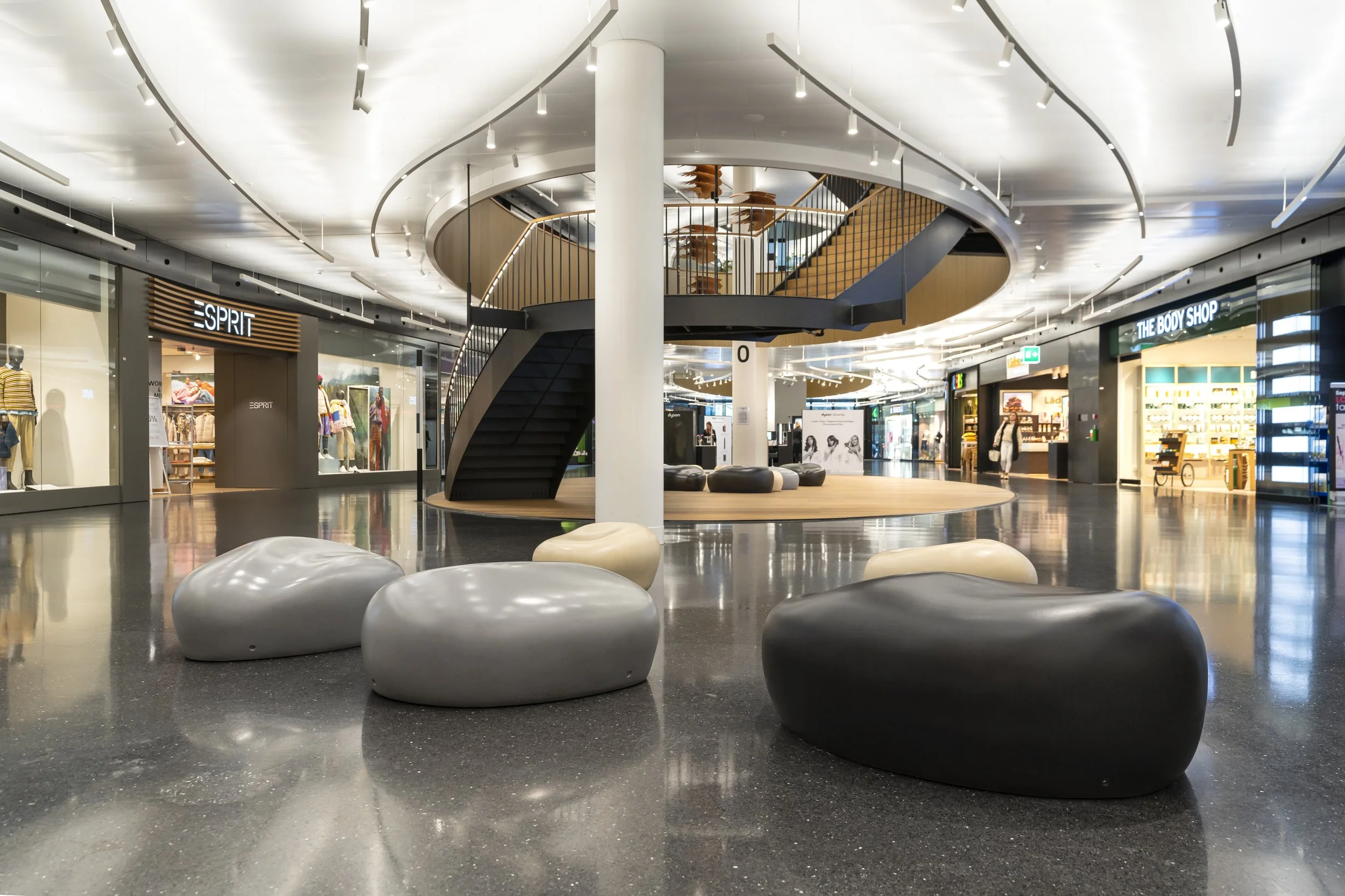 Leerer Einkaufst mall mit Sitzmöbeln aus Kunststoff in den Farben Schwarz, Grau und Weiß, mehrere Geschäfte im Hintergrund, eine Wendeltreppe führt zur oberen Etage, helle Beleuchtung und modernes Design.