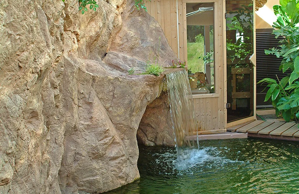 Ein künstlich angelegter Wasserfall fließt eine sandsteinfarbene Kunstfelsenformation herab  in einen Teich vor einer Zugangstür zu einer Sauna mit viel Grünpflanzen drum herum.