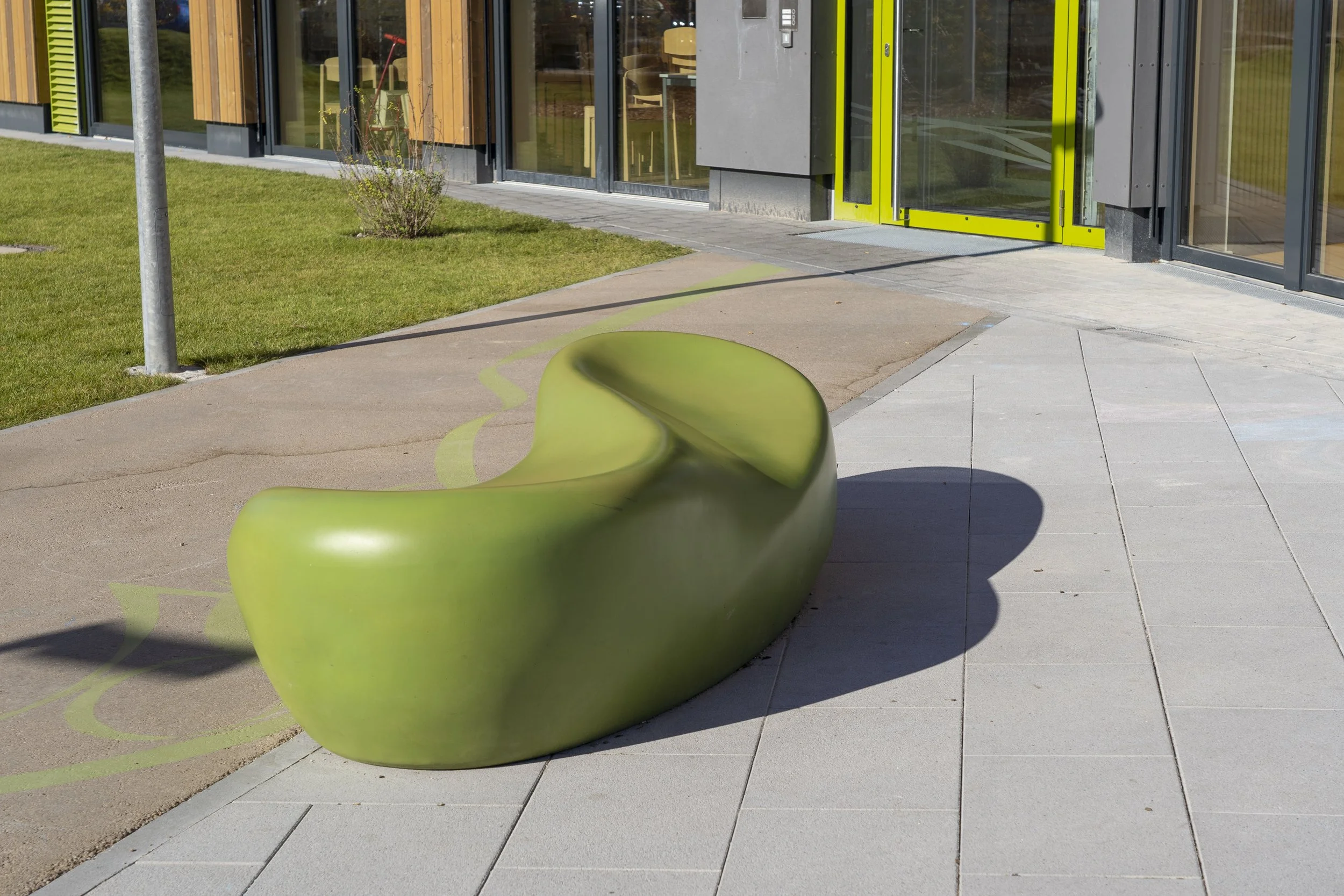 Moderne grüne Sitzbank vor einem Gebäude mit großen Fenstern und gelbgrünen Türen, umgeben von Gehweg und Rasen.