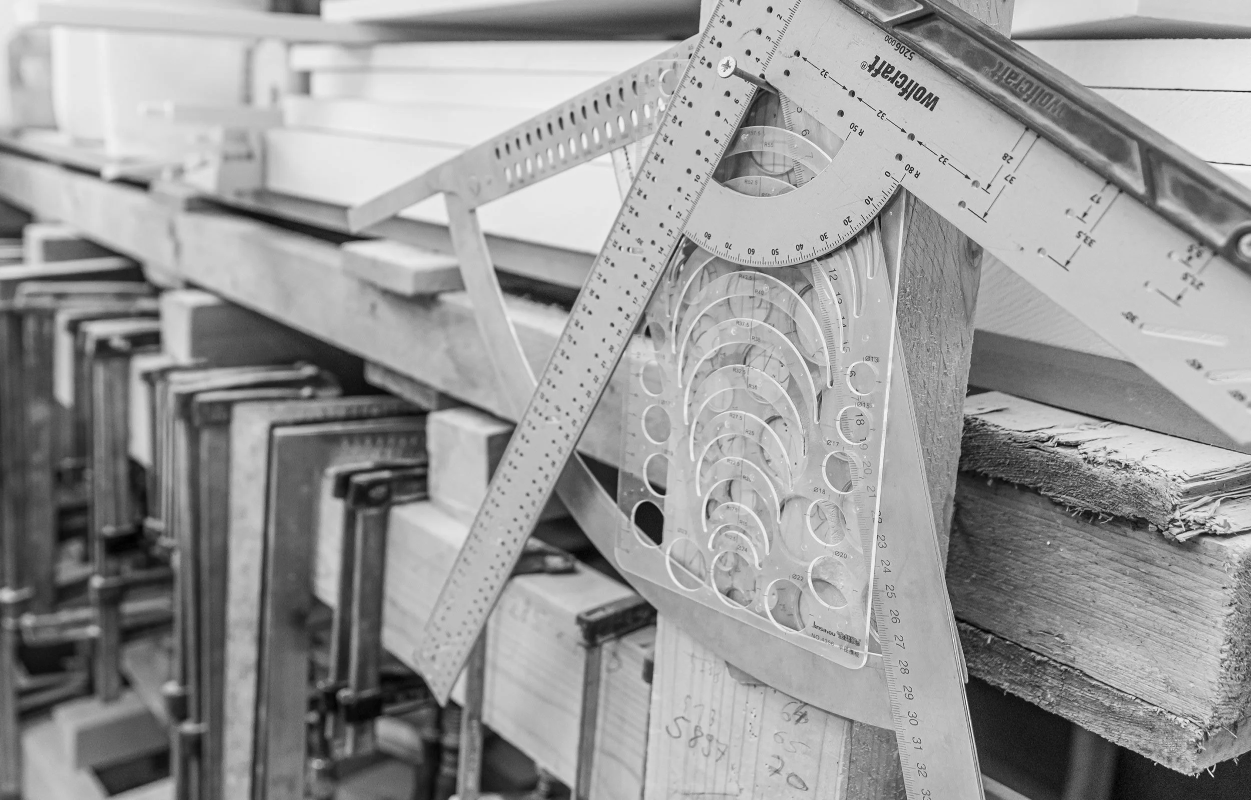 Ein Regal mit Schraubzwingen in verschiedenen Größen in einer Werkstatt in perspektivischer Ansicht. Im Vordergrund sind mehrere Winkelmesser und andere Messwerkzeuge zu sehen.