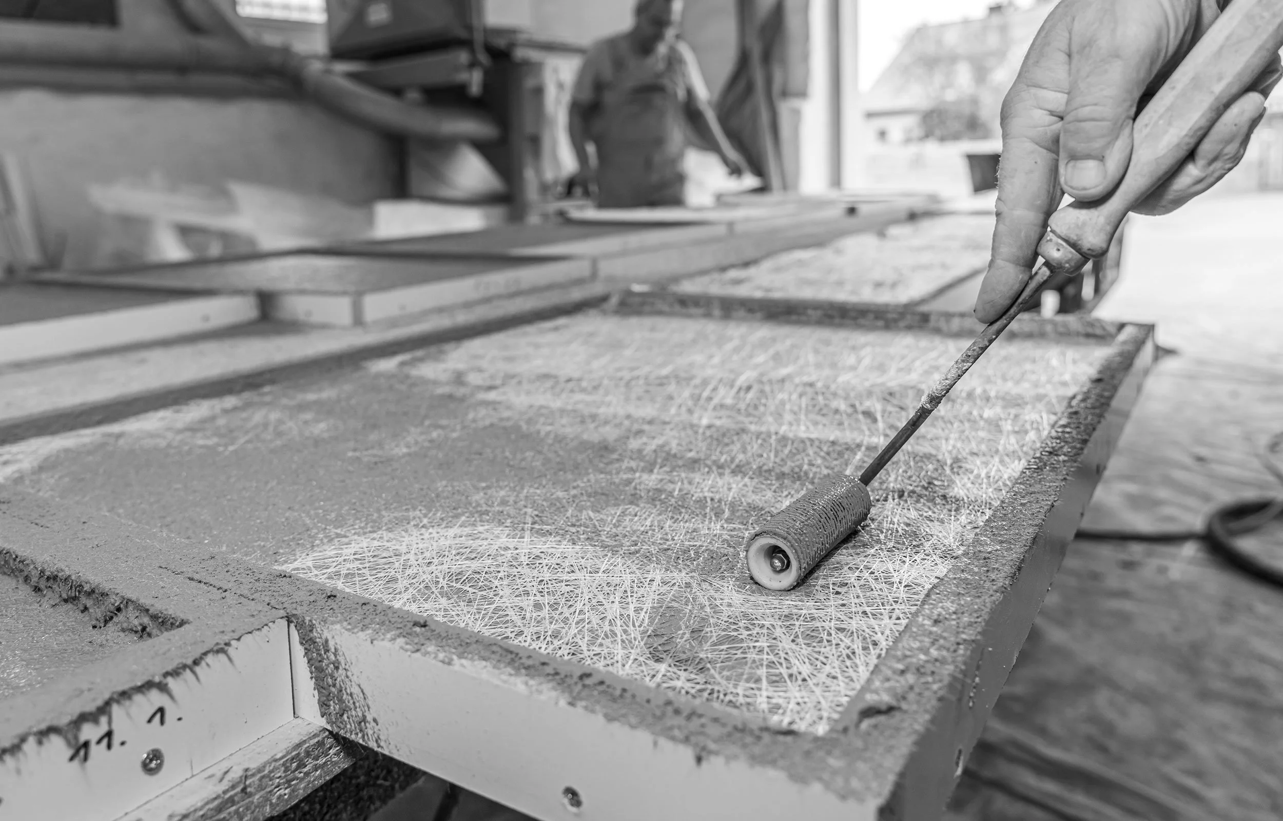 Bildausschnitt mit Nahaufnahme einer Hand, welche eine Gussform für Beton mit einem Farbroller bearbeitet, die Umgebung ist eine Werkstatt mit offenem Tor.