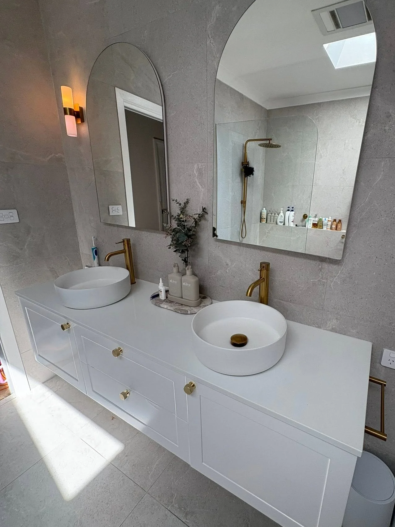 Modern bathroom with a white double vanity, round vessel sinks with gold faucets, large arched mirrors, wall-mounted light fixture, and a shower area with a gold showerhead visible in the mirror reflection.