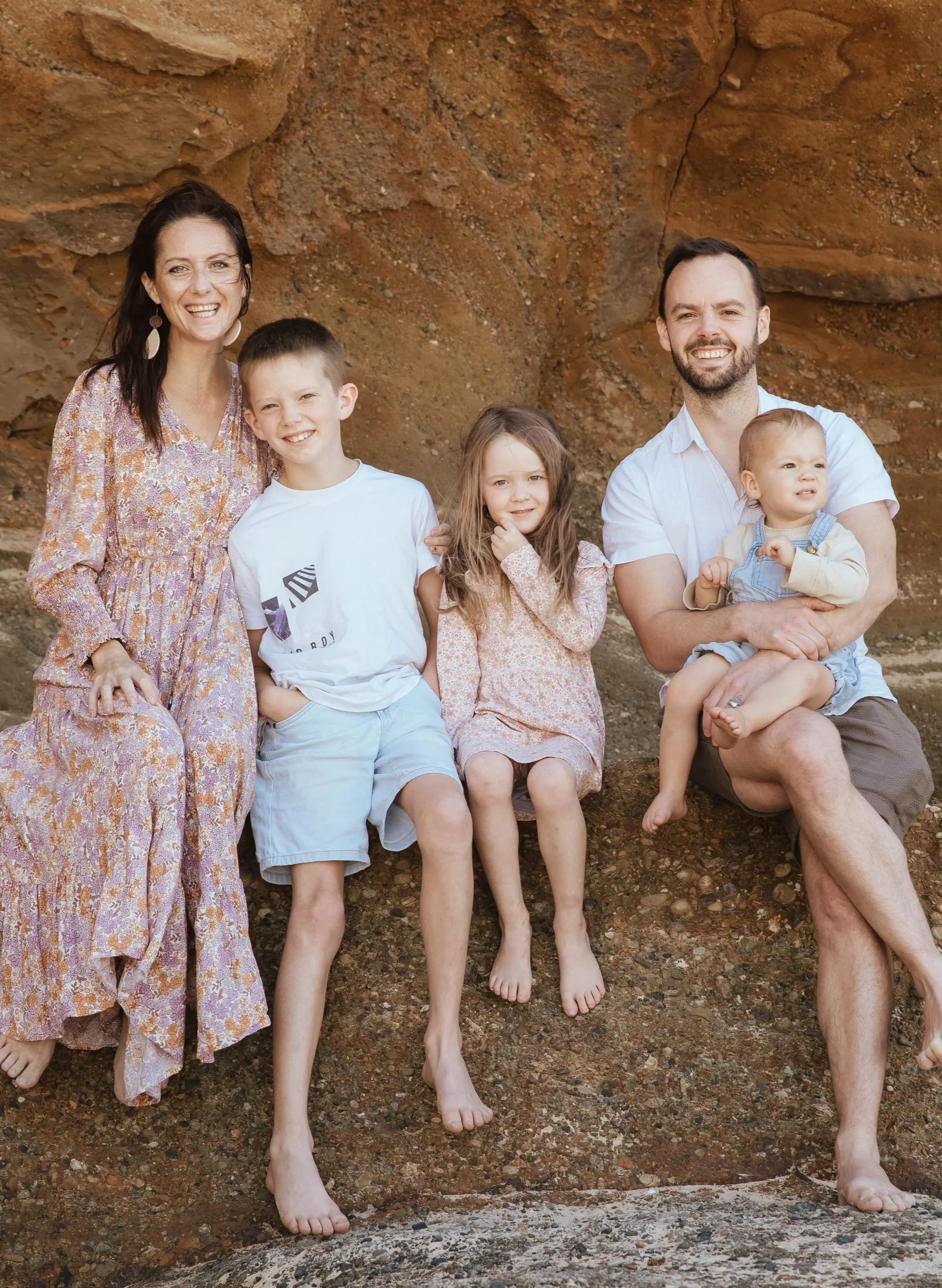 A family of six with light skin and brown hair sitting and standing in front of a rock wall, with only the bottom of their legs and feet visible.