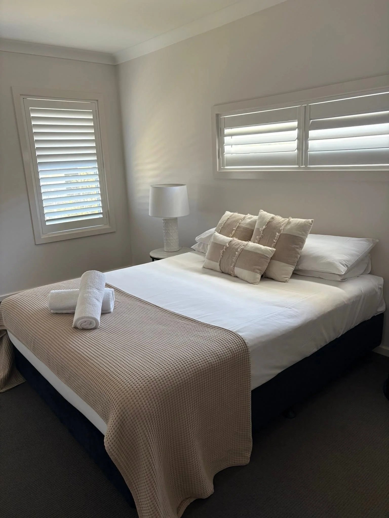 A bedroom with a neatly made bed, a beige checkered blanket at the foot, and white pillows with decorative beige pillows on top. There is a white nightstand with a white lamp near the bed. The room has two windows with white shutters, and the walls are painted white.