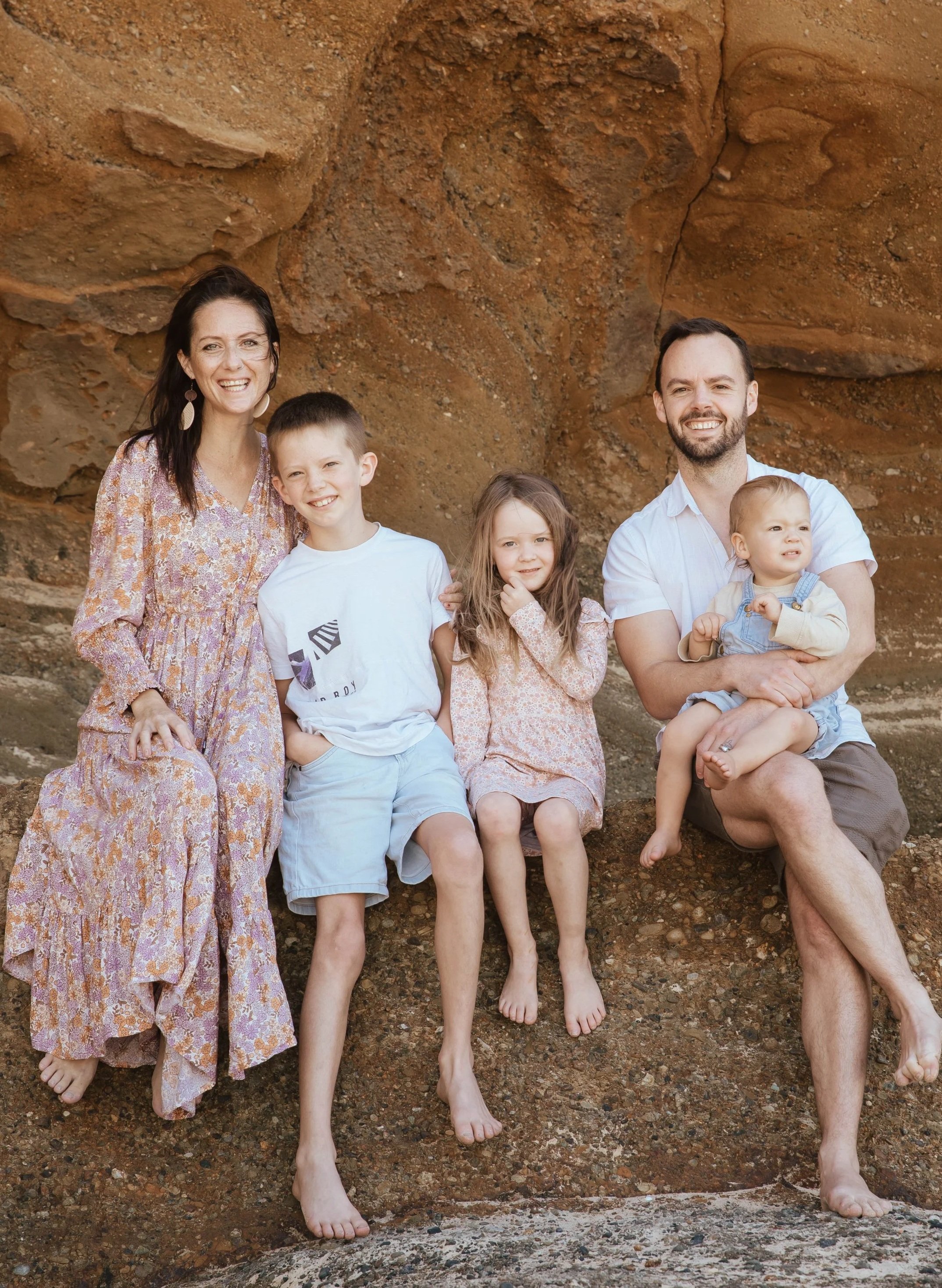 A family of five posing barefoot in front of a rocky, reddish-brown cliff. The group includes a woman, a man, and three children, all smiling and looking at the camera.