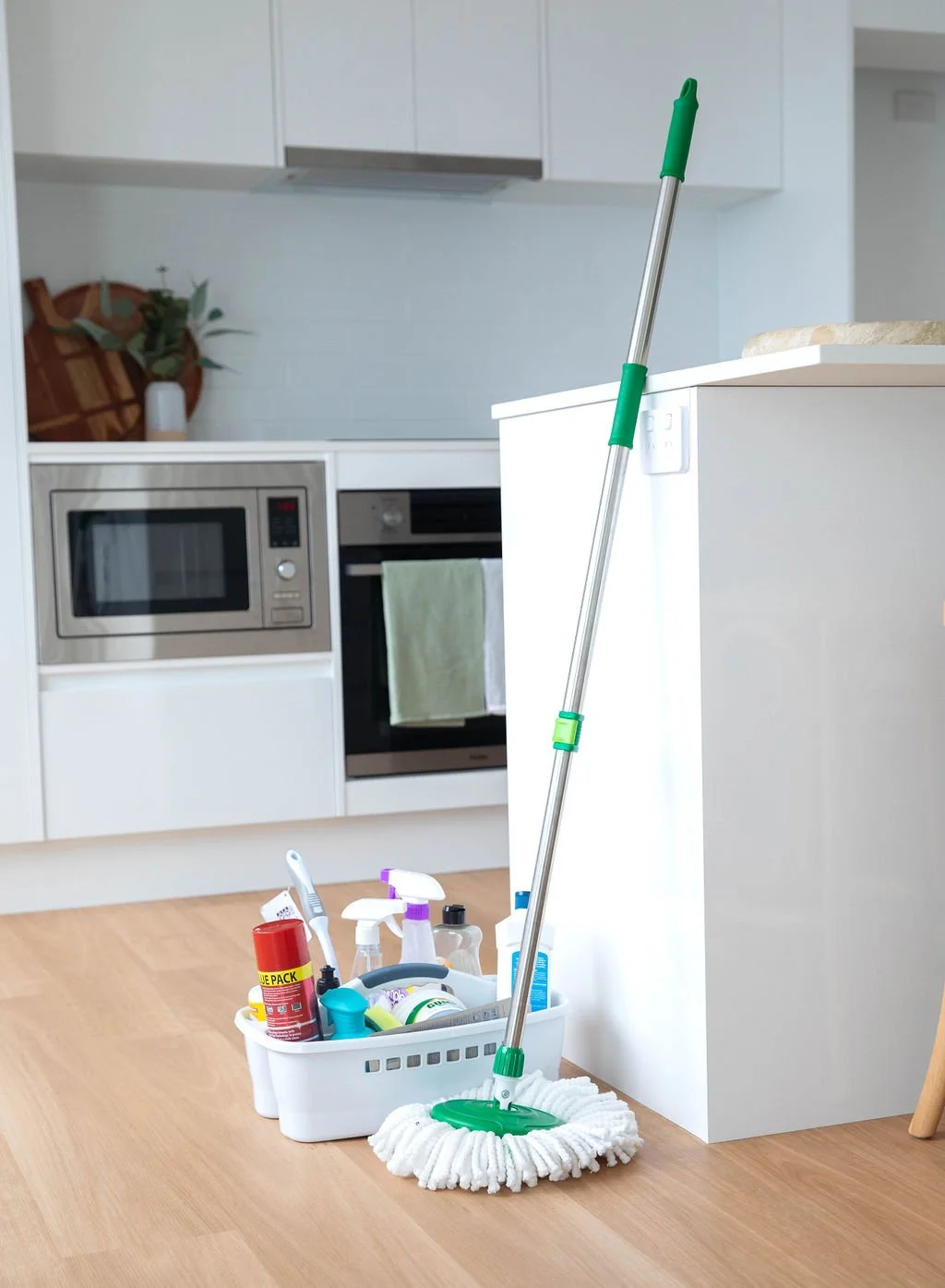 A mop with a green handle leaning against a white kitchen island, beside a white cleaning supplies basket on a wooden floor.