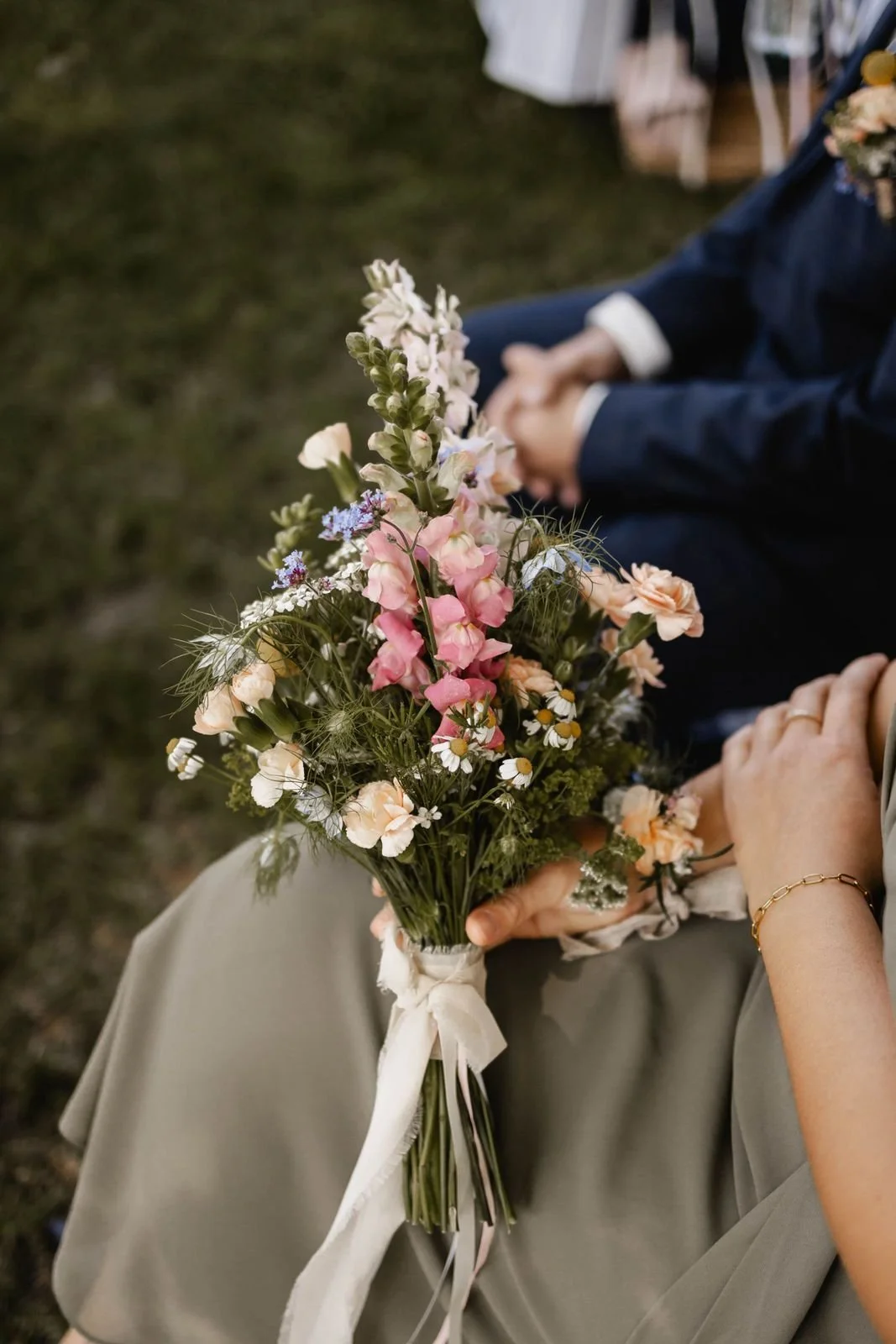 Nahaufnahme eines Brautstraußes mit verschiedenen Blumen in pastellfarben, gehalten von einer Person in einem Hochzeitskleid, bei einer Hochzeit im Freien.